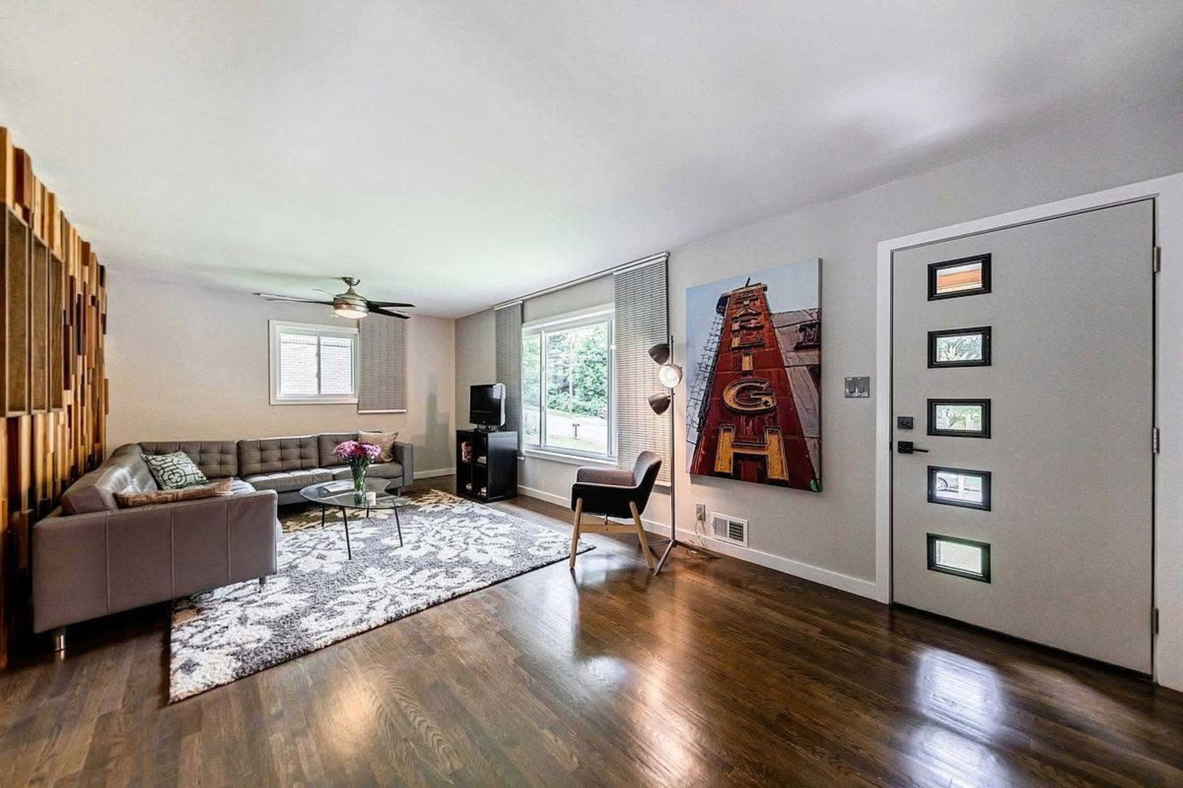 The image shows a modern living room featuring a gray sectional sofa, a round glass coffee table, and a large window that lets in natural light, with a door and a distinctive piece of wall art.
