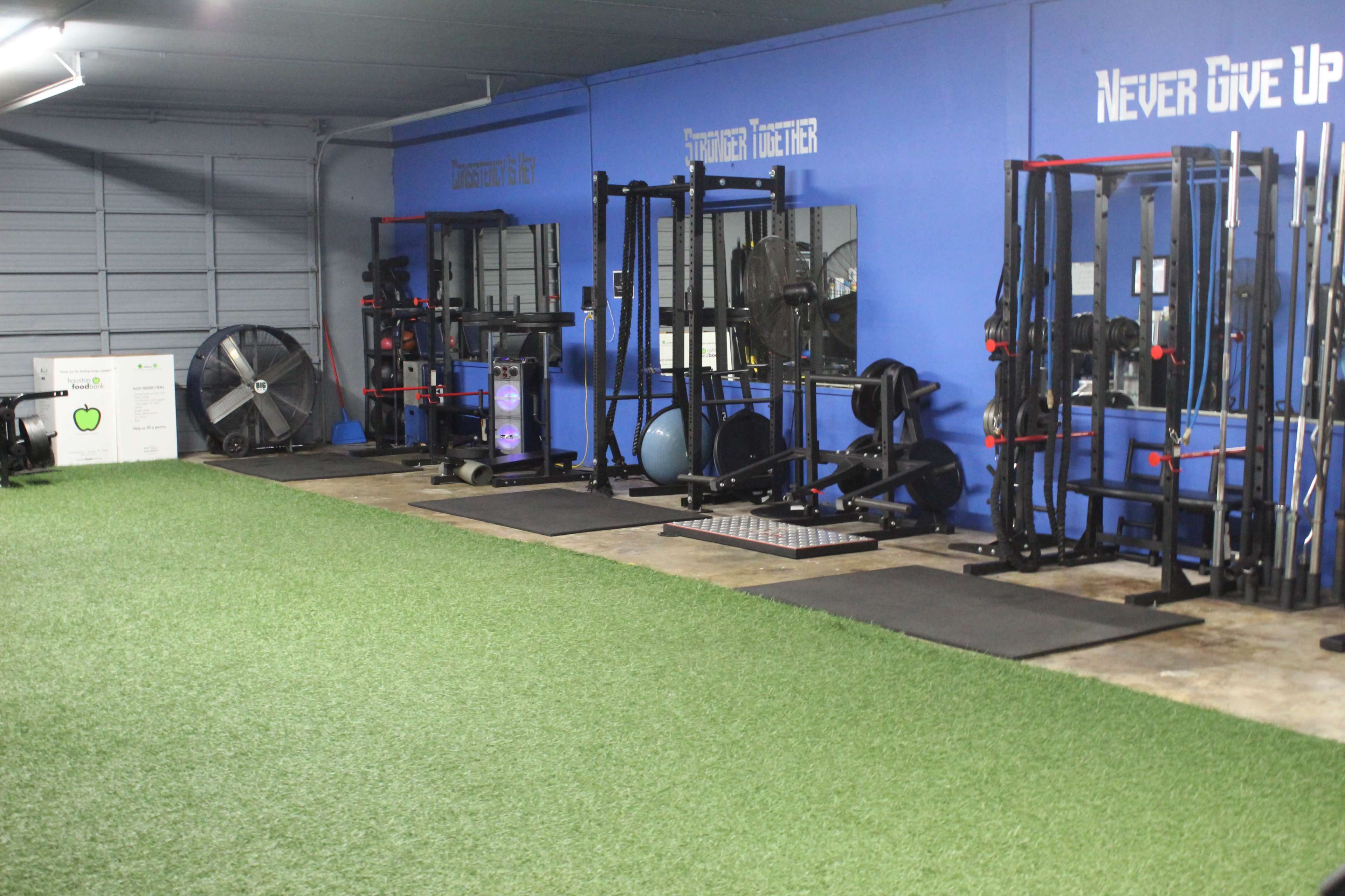 A well-equipped gym with various exercise machines, weights, and turf flooring along one side of a blue wall.