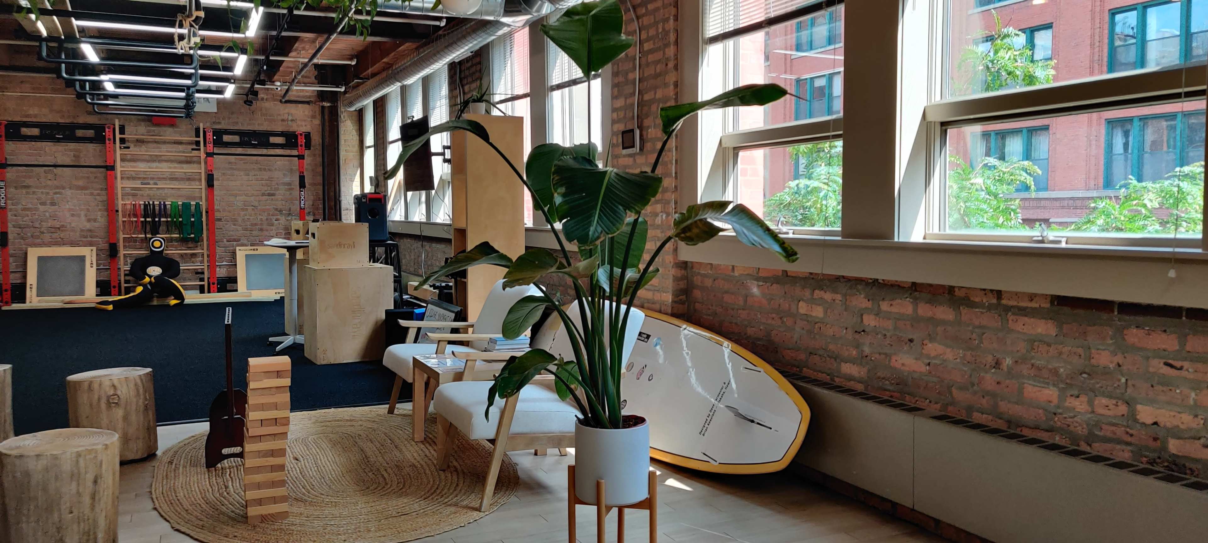 A spacious room with exposed brick walls, featuring a potted plant, wooden furniture, exercise equipment, and a stand-up paddleboard leaning against the wall.