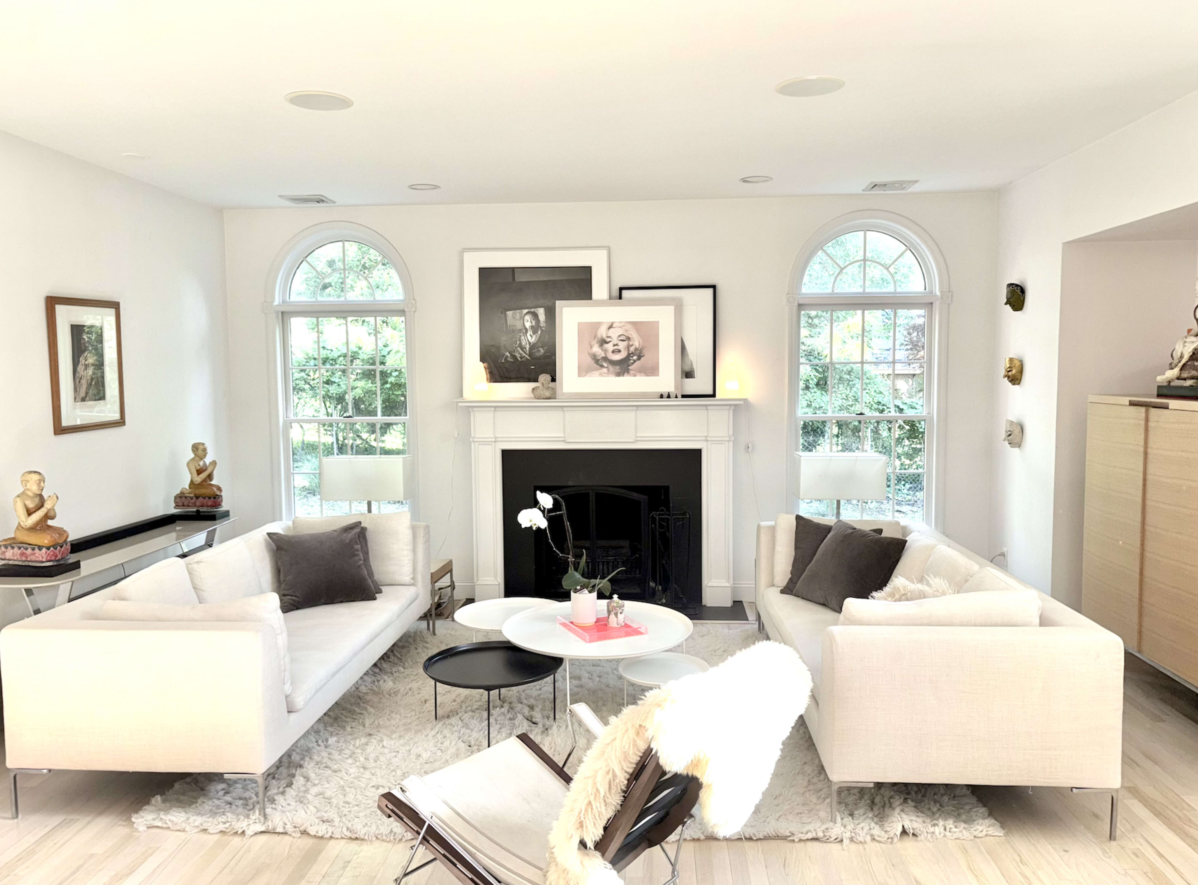 A bright living room features two white sofas facing a small circular table, with large windows allowing natural light and displaying lush greenery outside.
