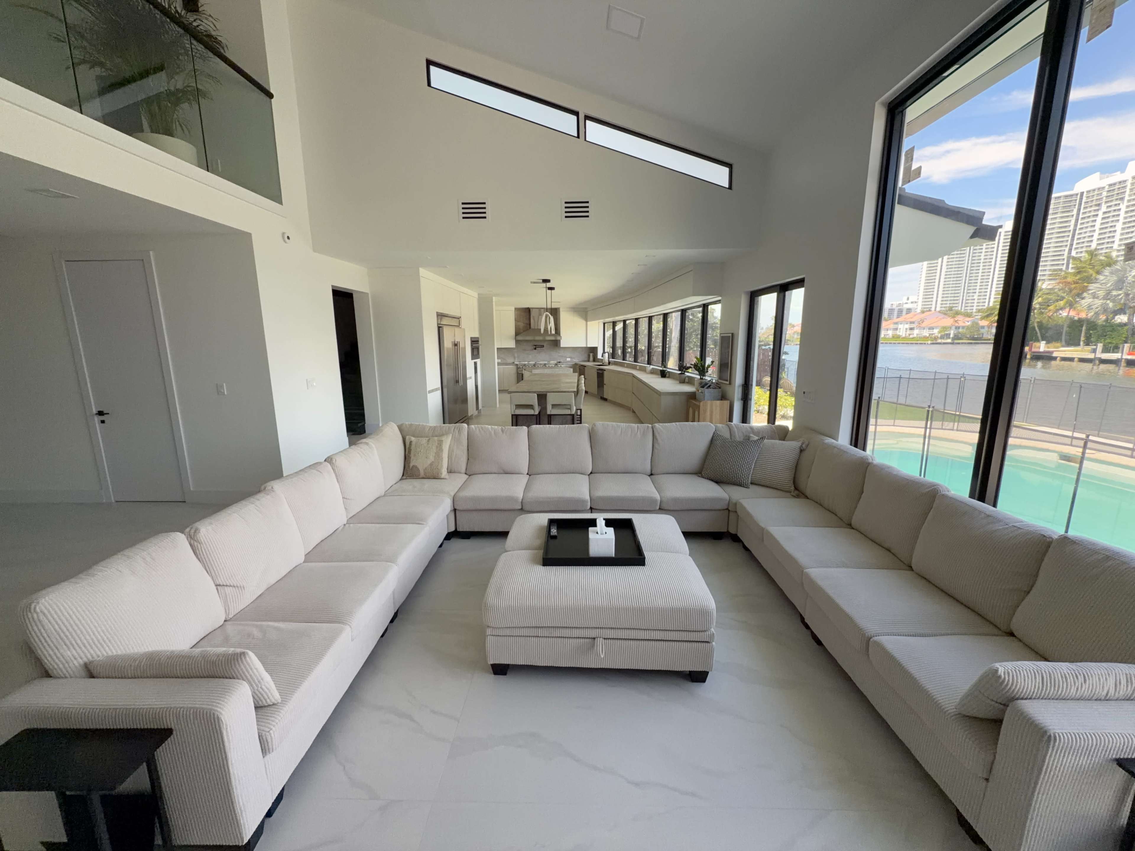 The image shows a spacious, modern living room with a large U-shaped sectional sofa and a coffee table, overlooking a pool area through expansive windows.