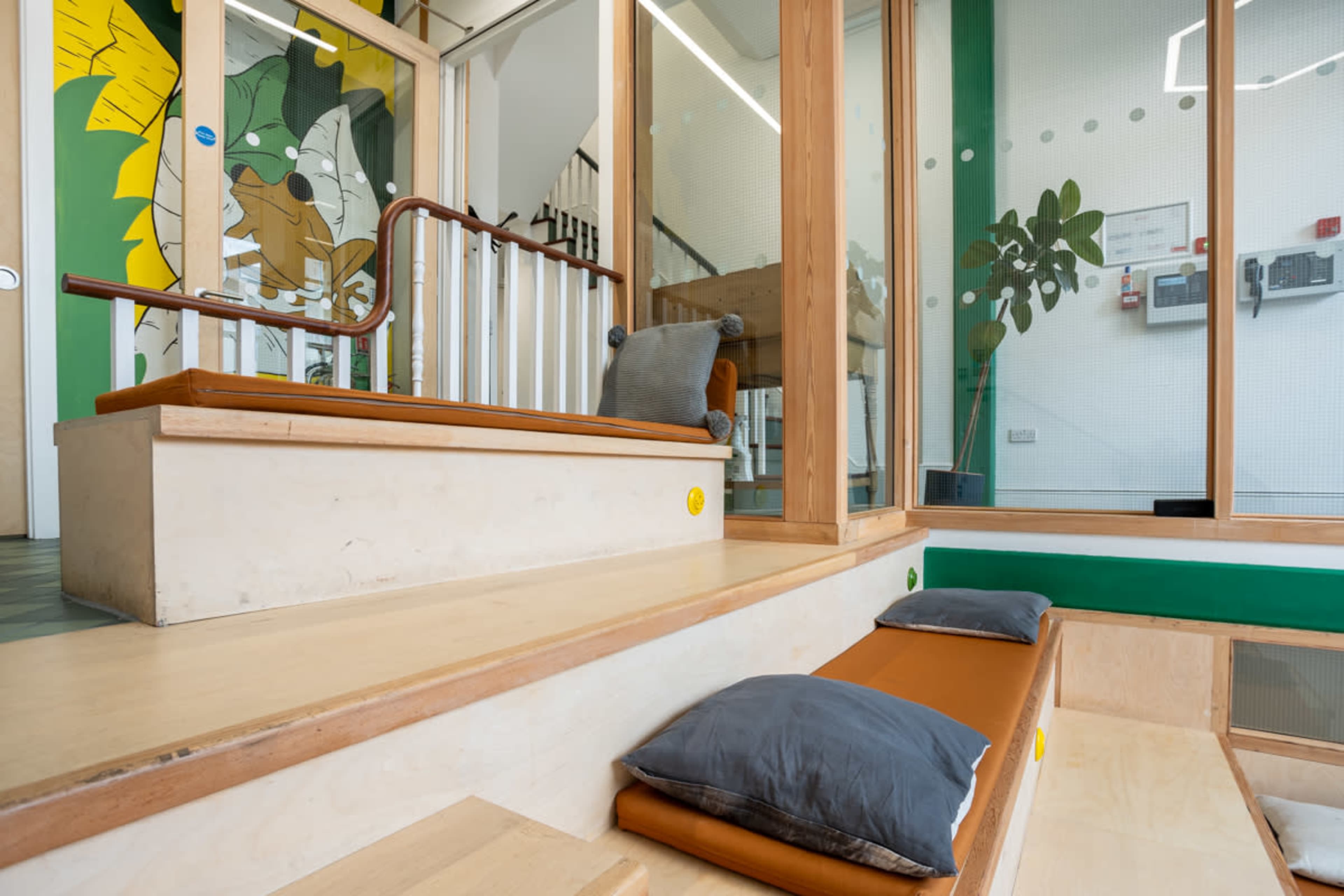 A bright indoor space with wooden steps, cushions, and a glass partition featuring a green mural of a cartoon animal.