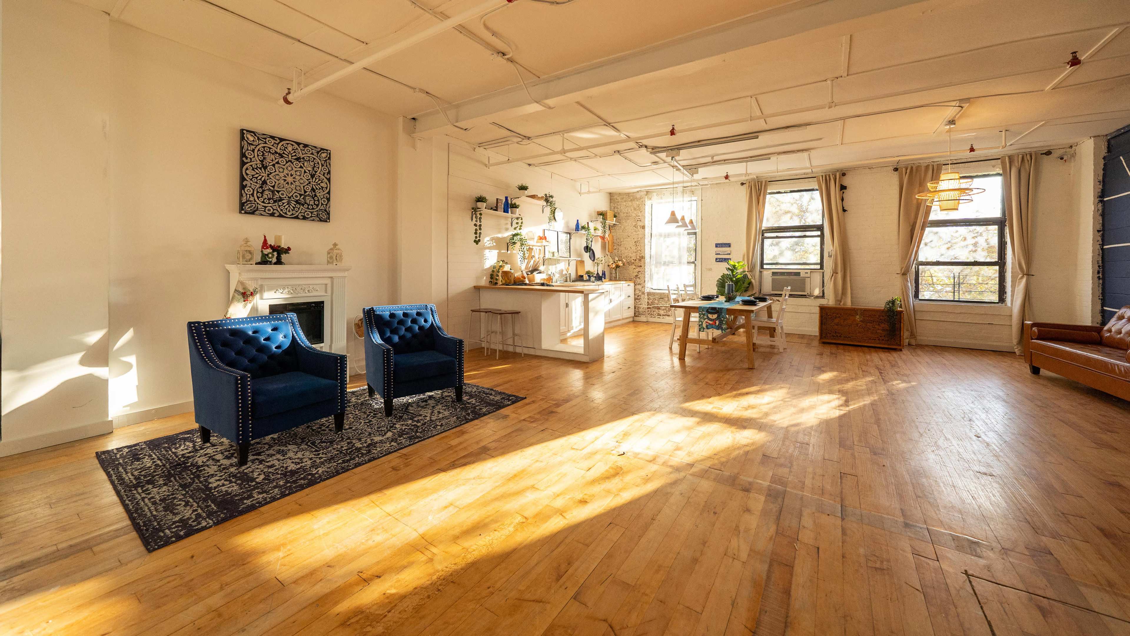 A spacious, well-lit room with wooden flooring, two blue chairs, and a kitchen area in the background.