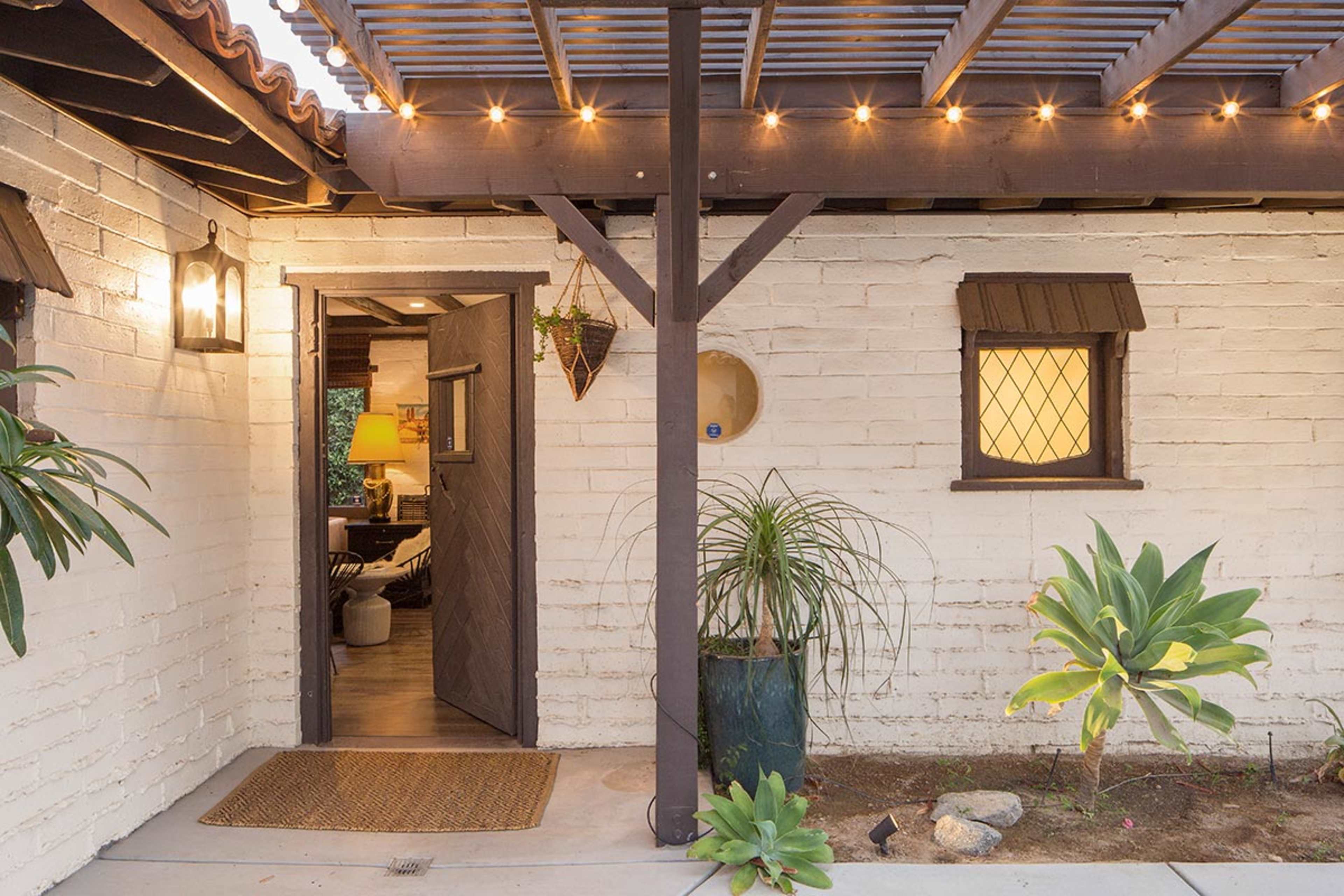 A doorway with a slatted overhang, leading to a warmly lit interior, surrounded by potted plants and string lights.