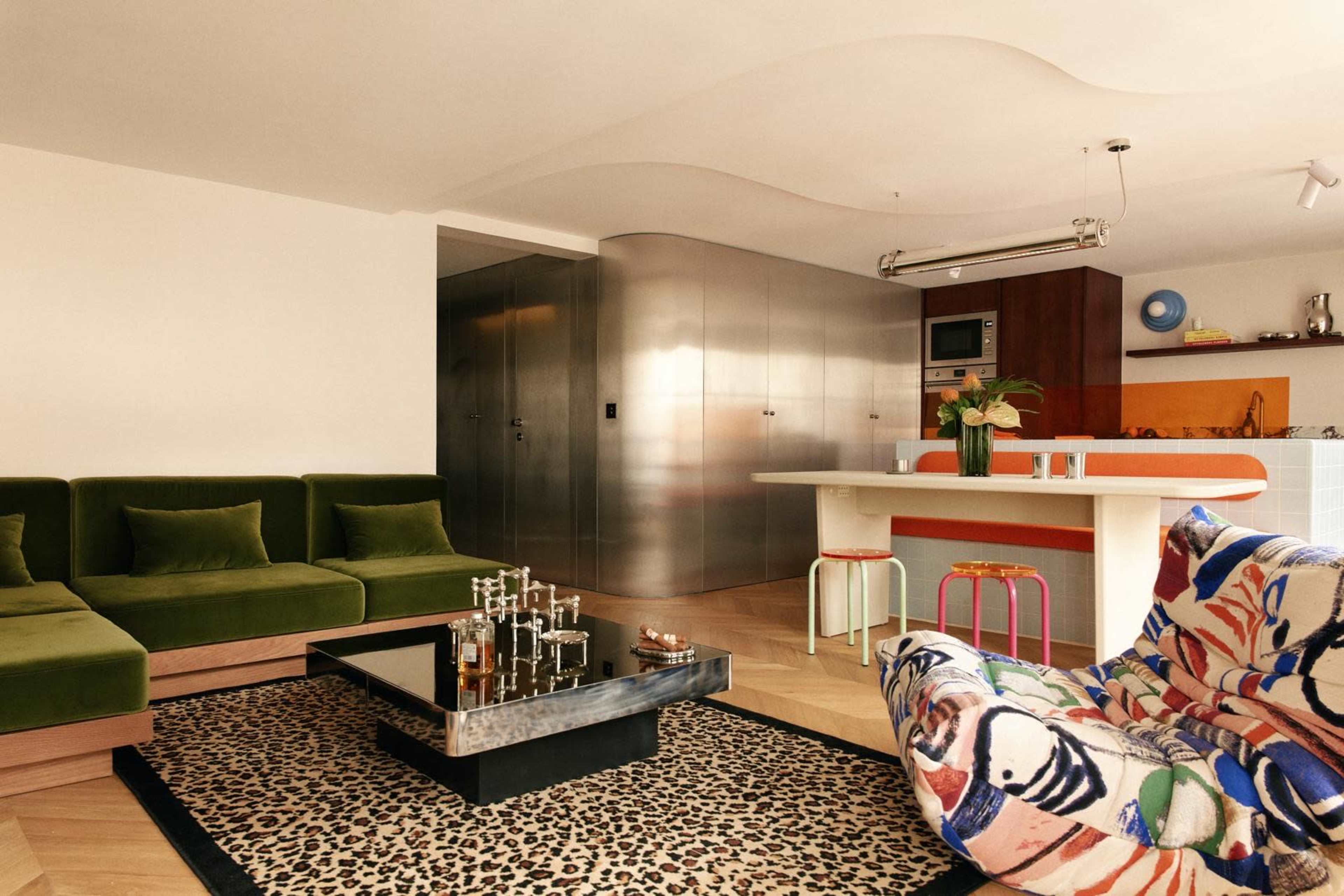 The image shows a modern living space featuring a green sectional sofa, a leopard print rug, a glass coffee table, and a kitchen area with stainless steel cabinets.