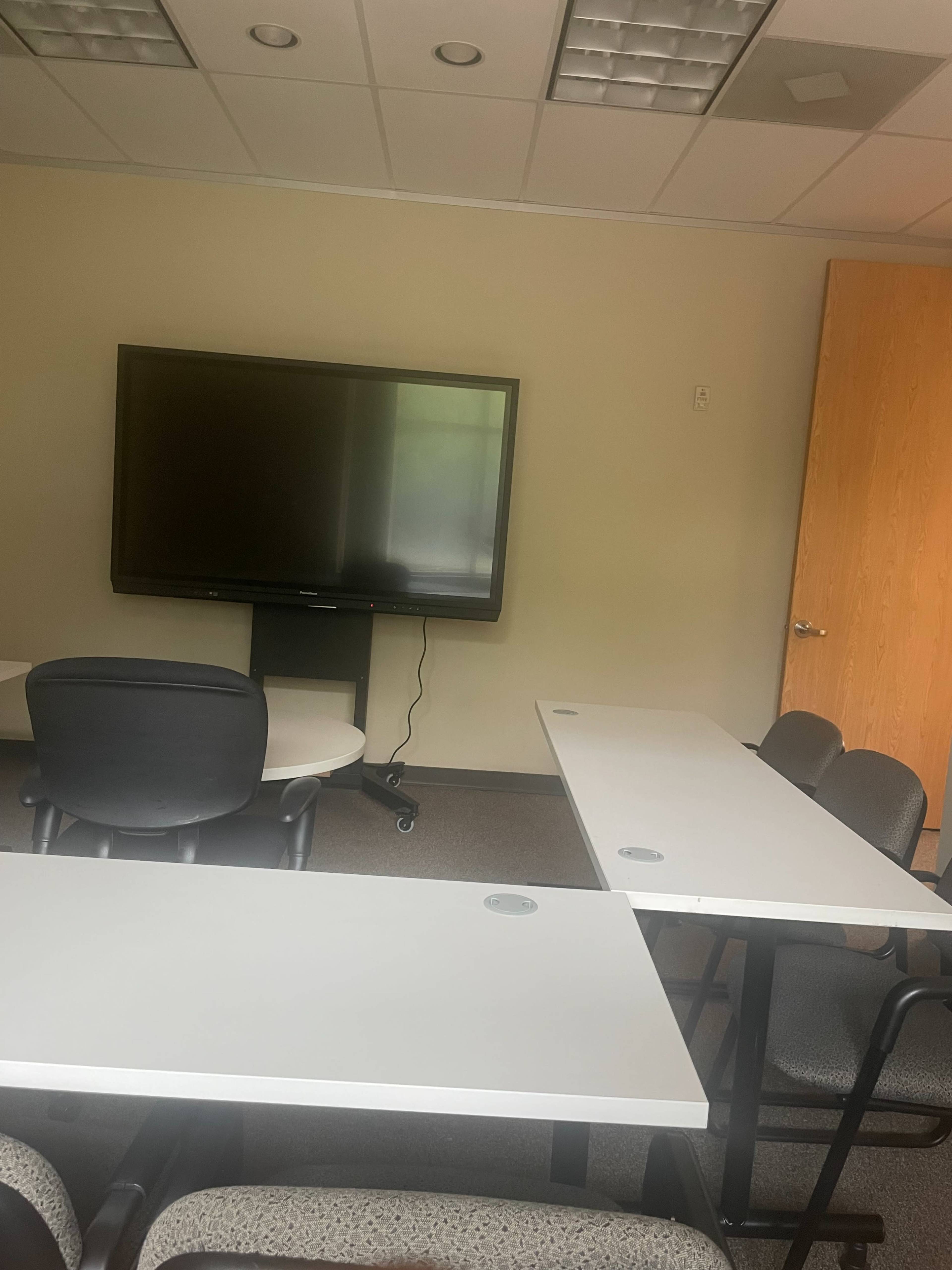 A meeting room with a large screen, a round table, and several chairs arranged around rectangular tables.