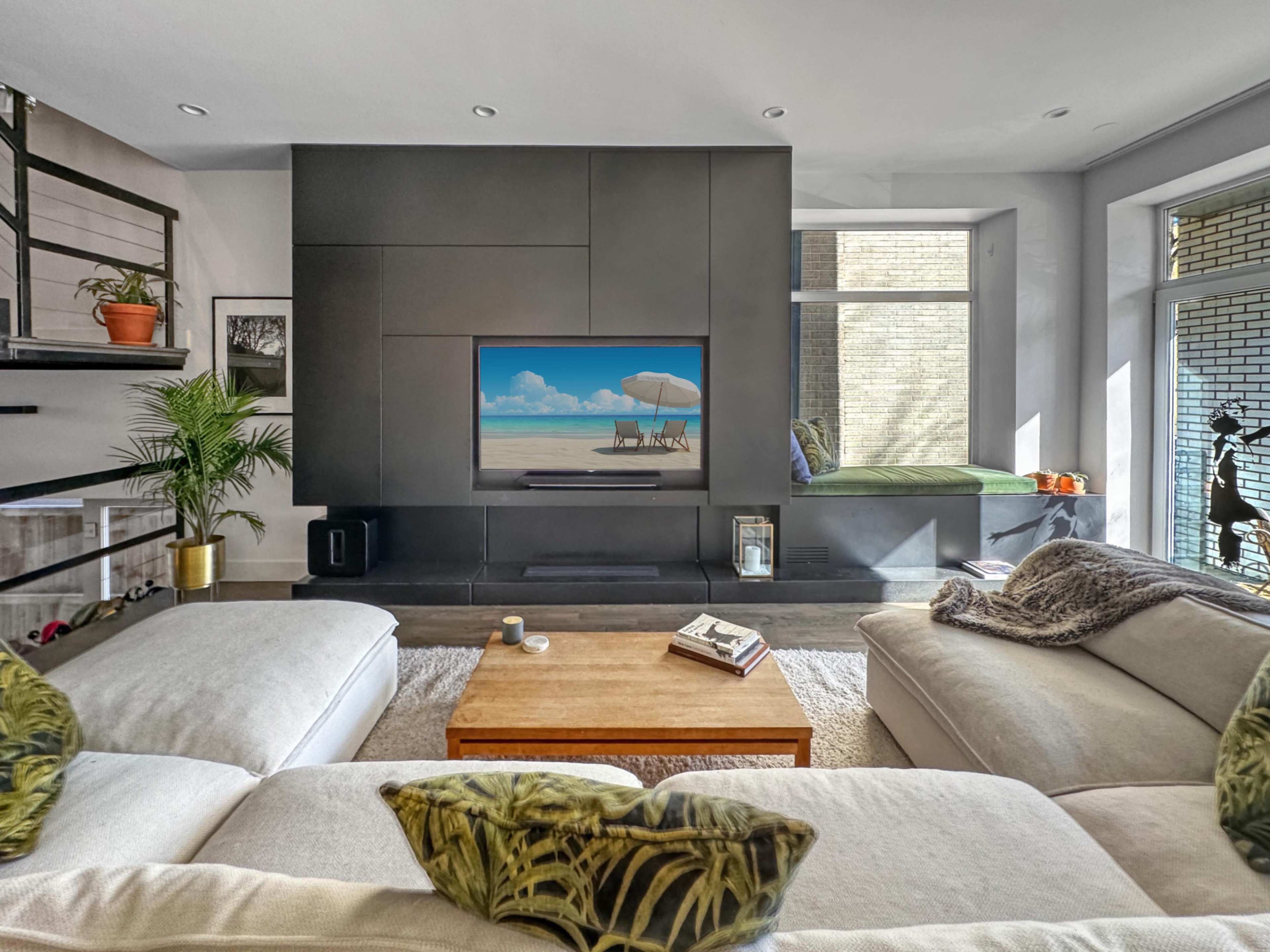 A modern living room features a large sectional sofa, a wooden coffee table, and a wall-mounted TV displaying a beach scene, with a sunny window seat visible in the background.