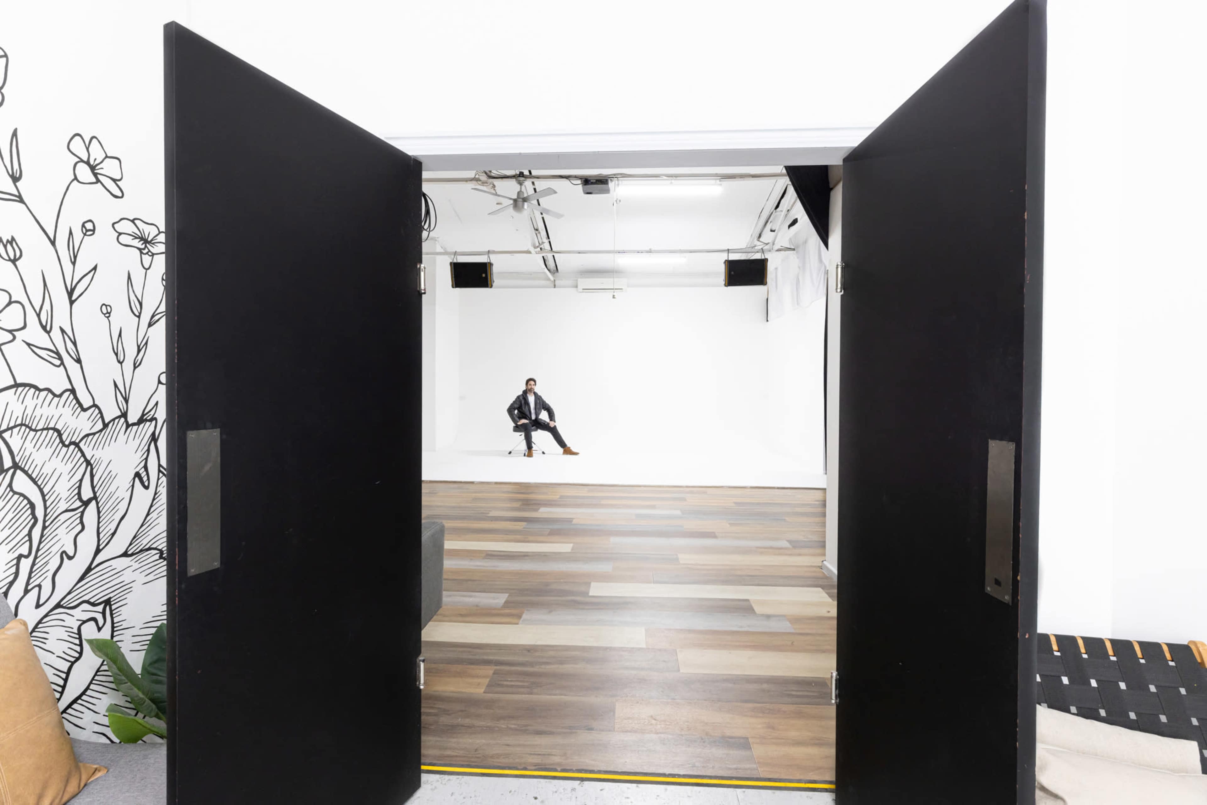 A person sits on a stool in a well-lit, empty studio, framed by large black doors that are wide open.