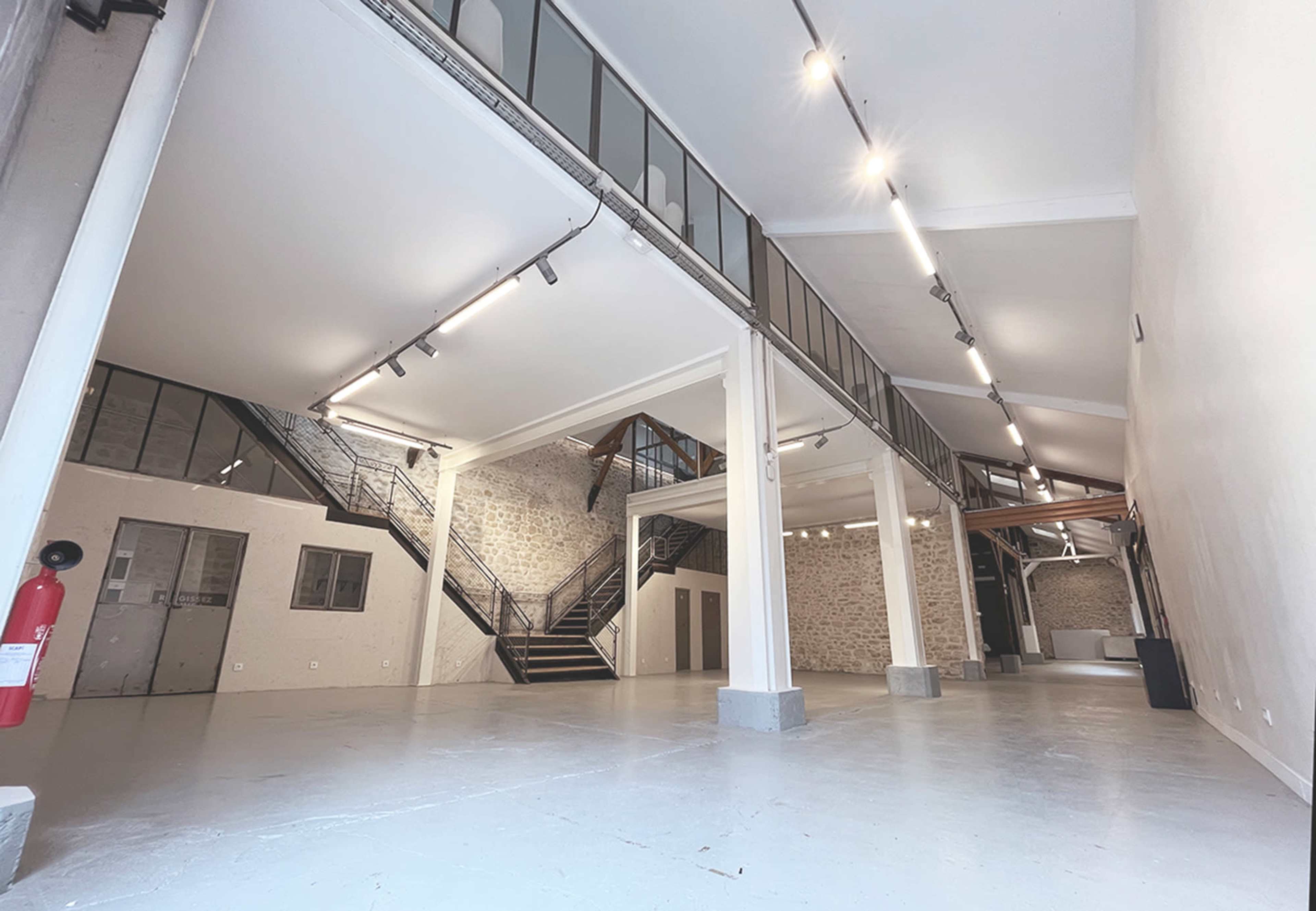 An open, well-lit industrial space with high ceilings, featuring exposed brick walls, a staircase, and minimalist design elements.