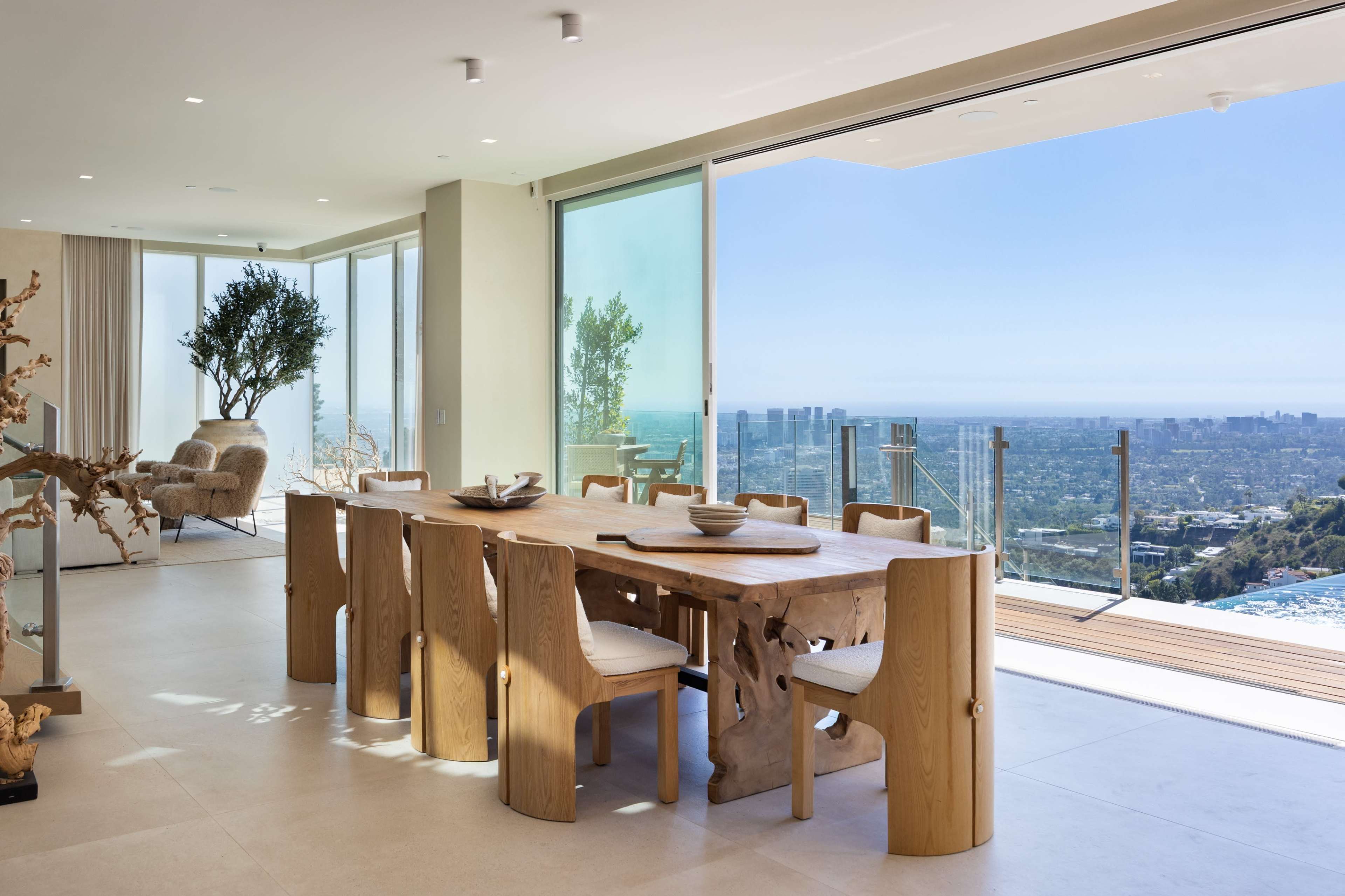 A spacious dining area features a large wooden table surrounded by uniquely designed chairs, with large glass doors offering a view of the cityscape and mountains beyond.