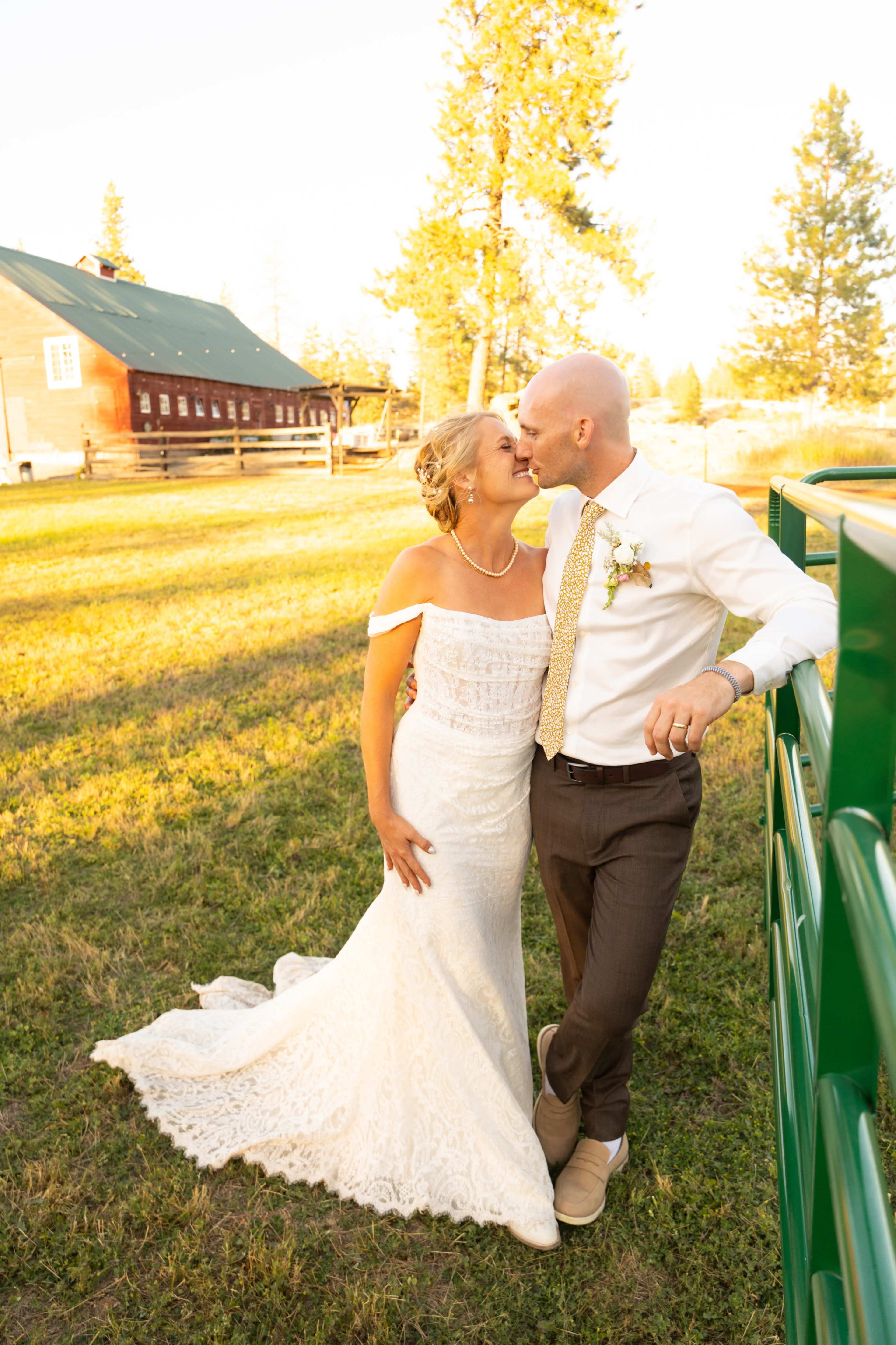 A bride and groom share a kiss in a sunlit outdoor setting, with a rustic barn and trees in the background.