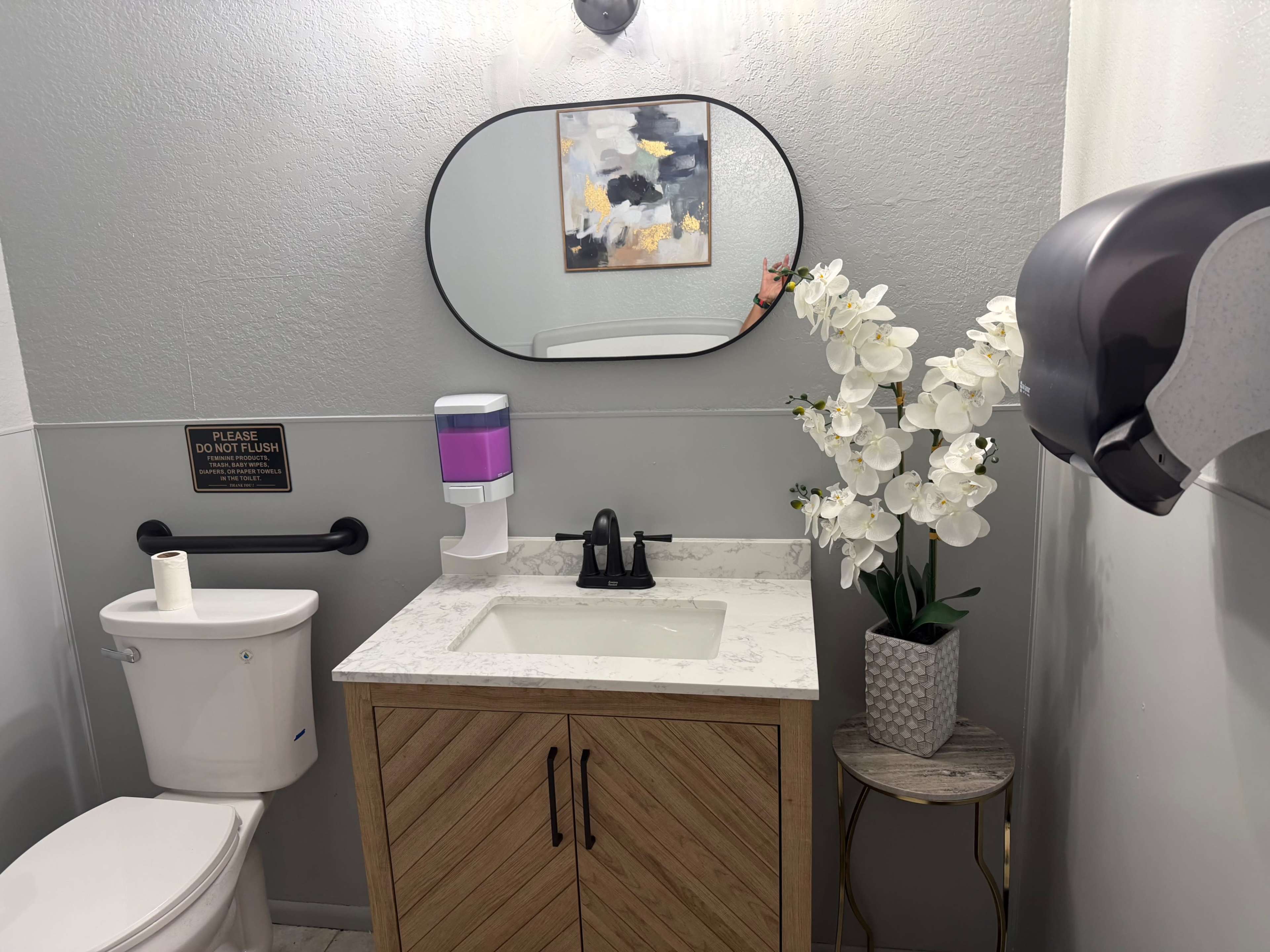 The image shows a modern bathroom with a toilet, a sink with a marble countertop, a decorative mirror, and a plant arrangement.
