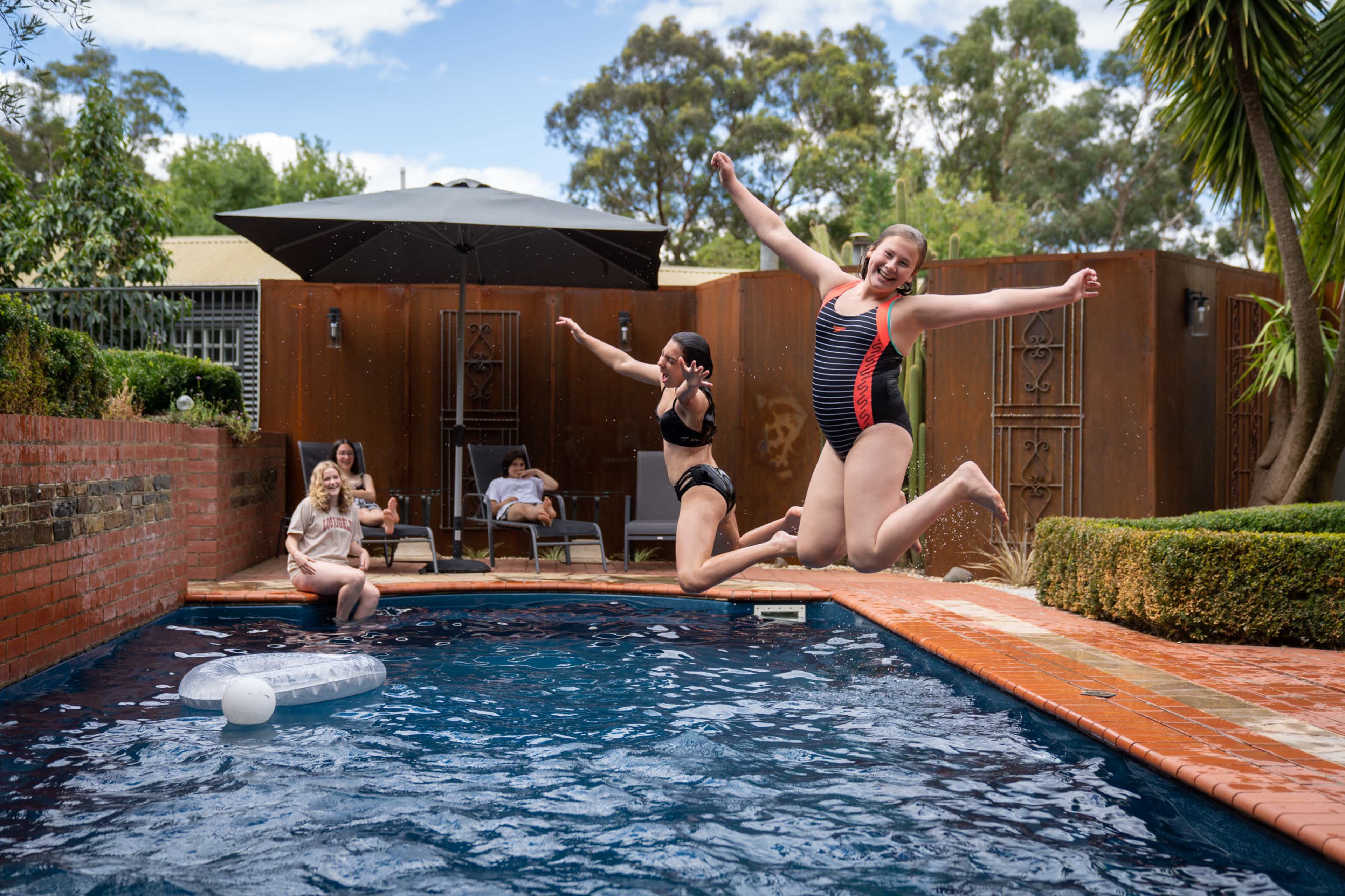 Two girls jump into a swimming pool while two others relax nearby on lounge chairs.