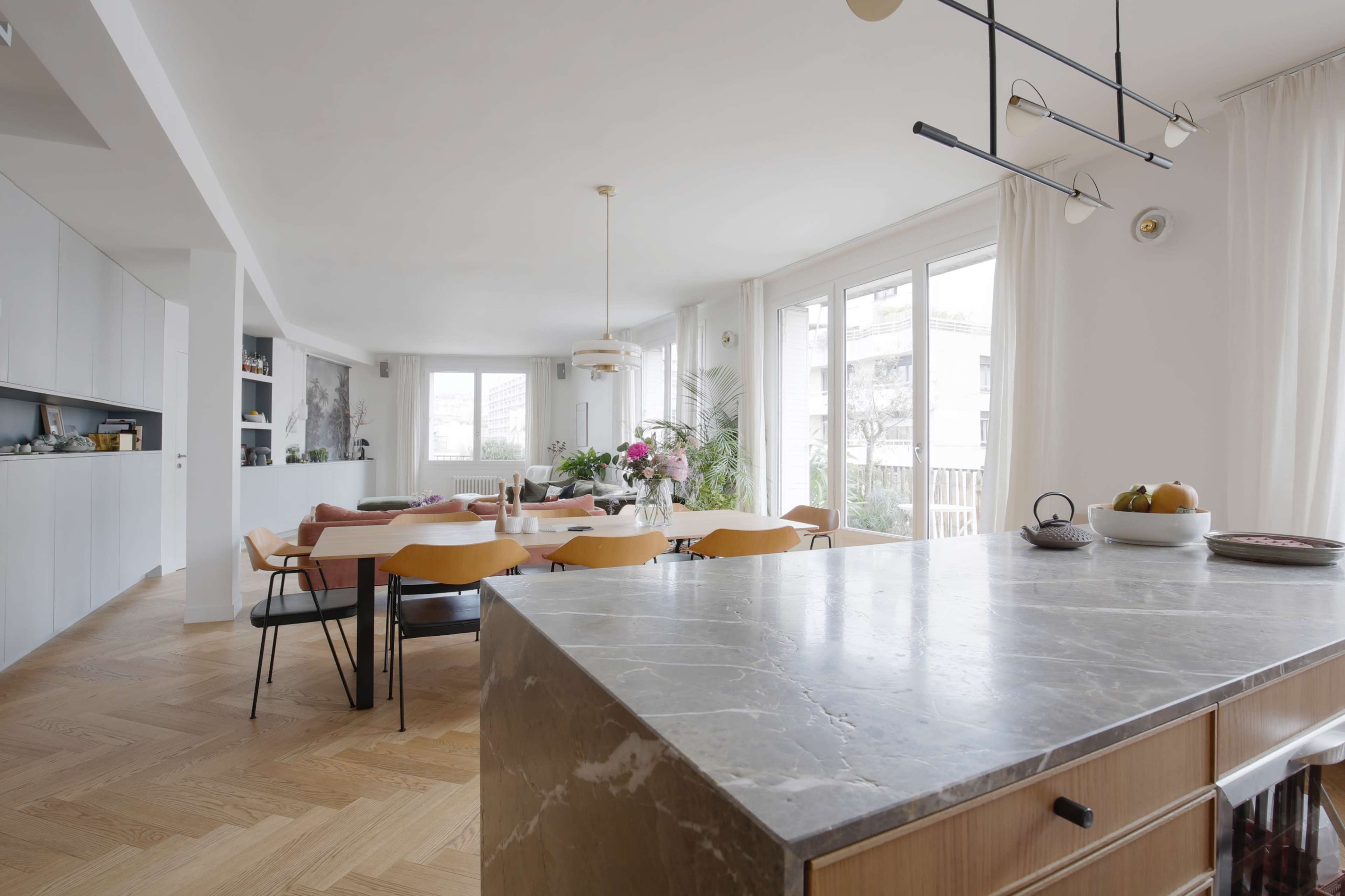 A modern open-concept living and dining area features a marble kitchen island, wooden dining table, and large windows with natural light.