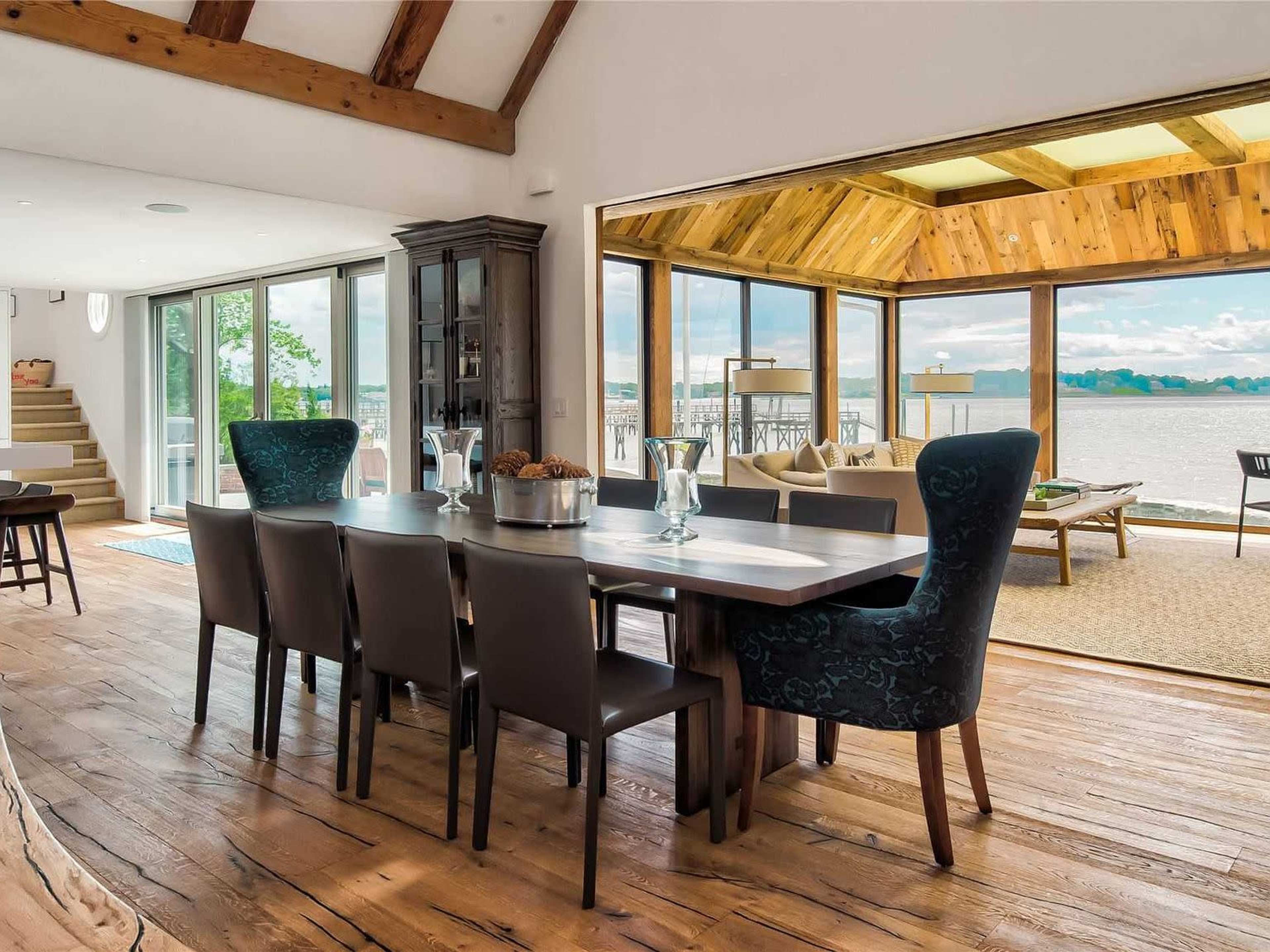 The image shows a spacious dining area with a large wooden table surrounded by chairs, connected to a sunlit room featuring large windows overlooking a waterfront view.