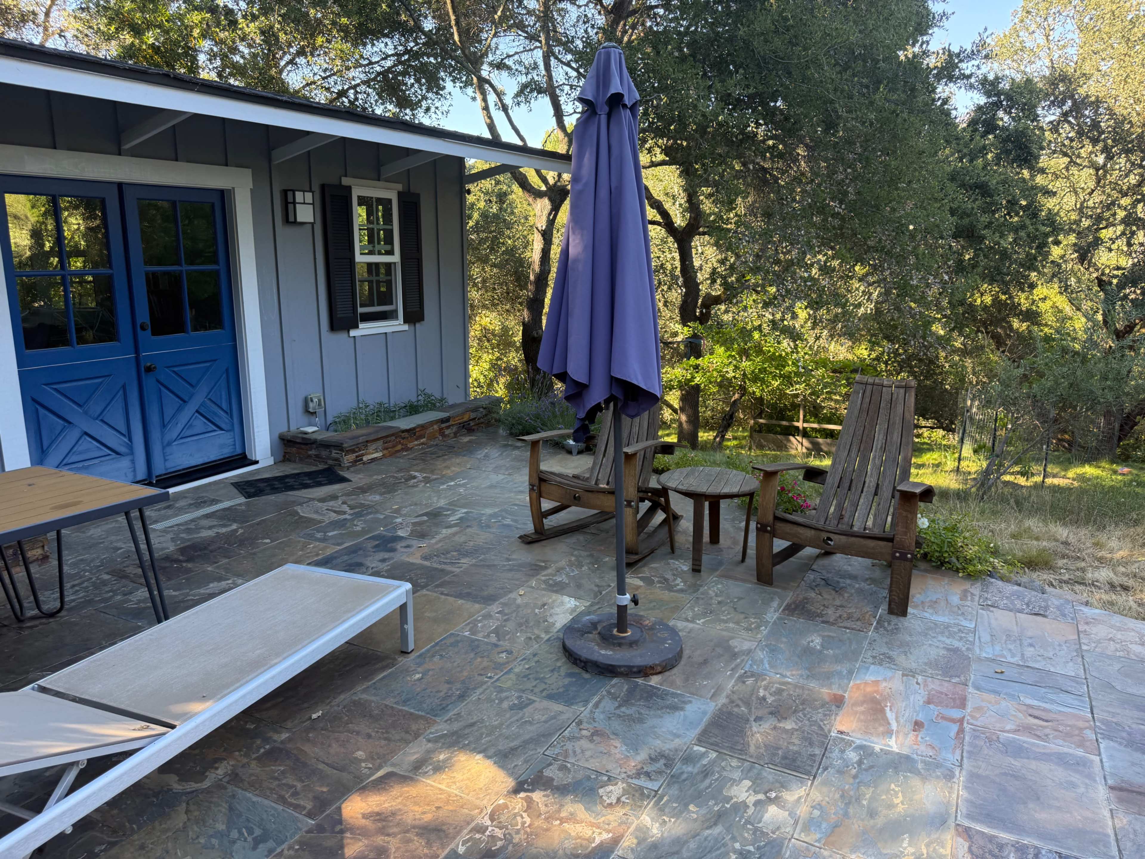 A patio area features a table, two wooden chairs, and a sun umbrella, surrounded by greenery and a blue door leading to a house.