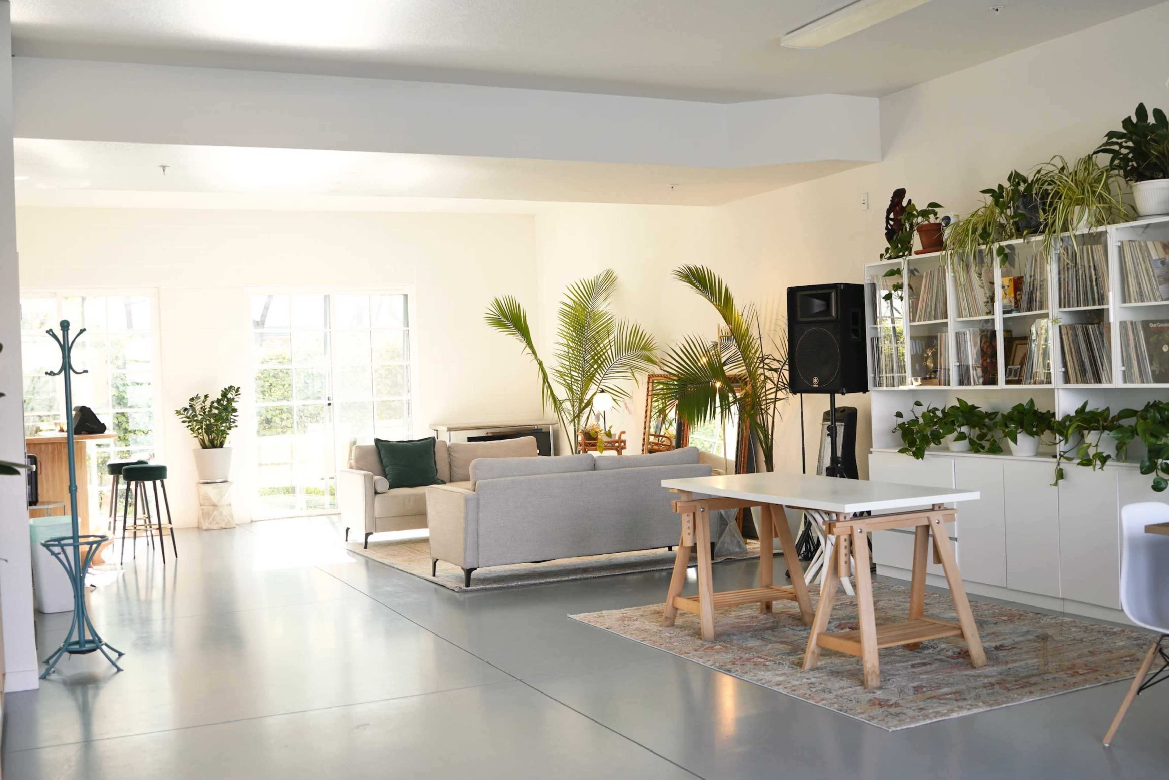 A bright, modern interior space featuring a living area with a sofa and plants, a dining table in the center, and shelves with records and greenery against a white wall.