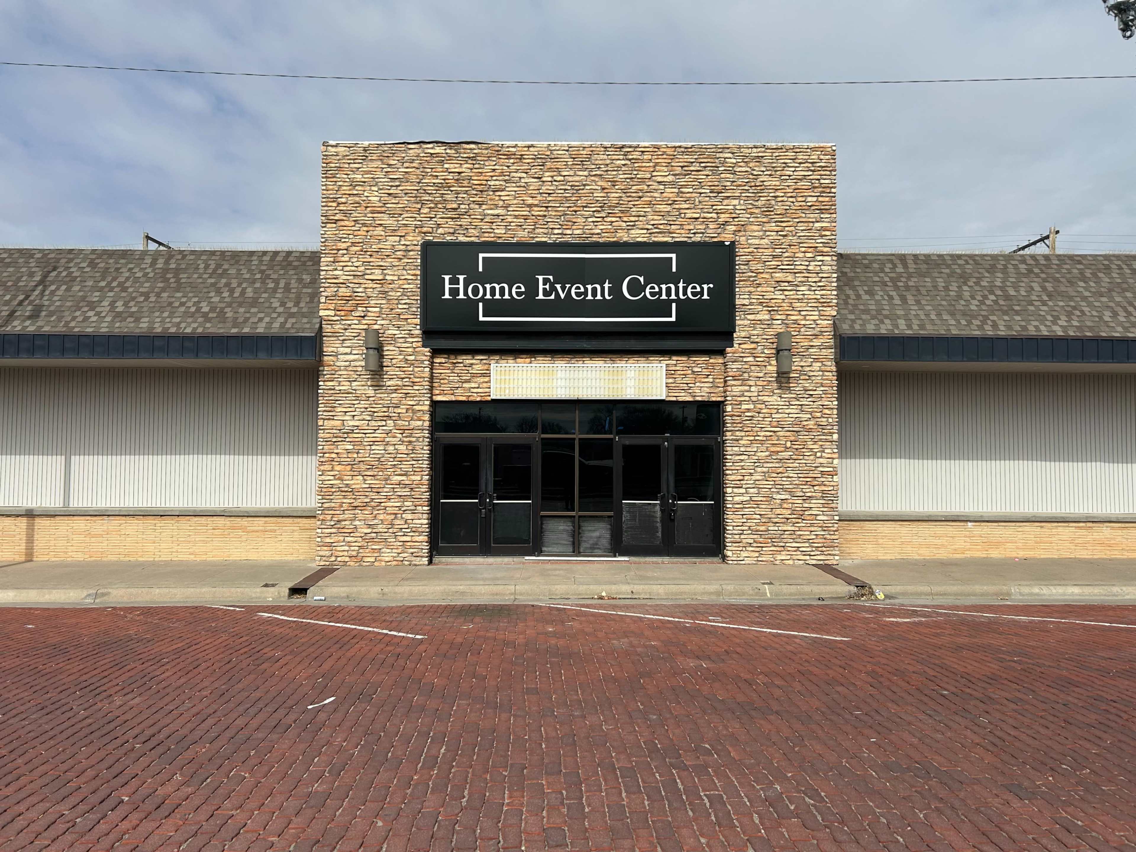 The image shows the front exterior of a building labeled "Home Event Center," featuring a stone facade and glass double doors, set against a brick-paved street.