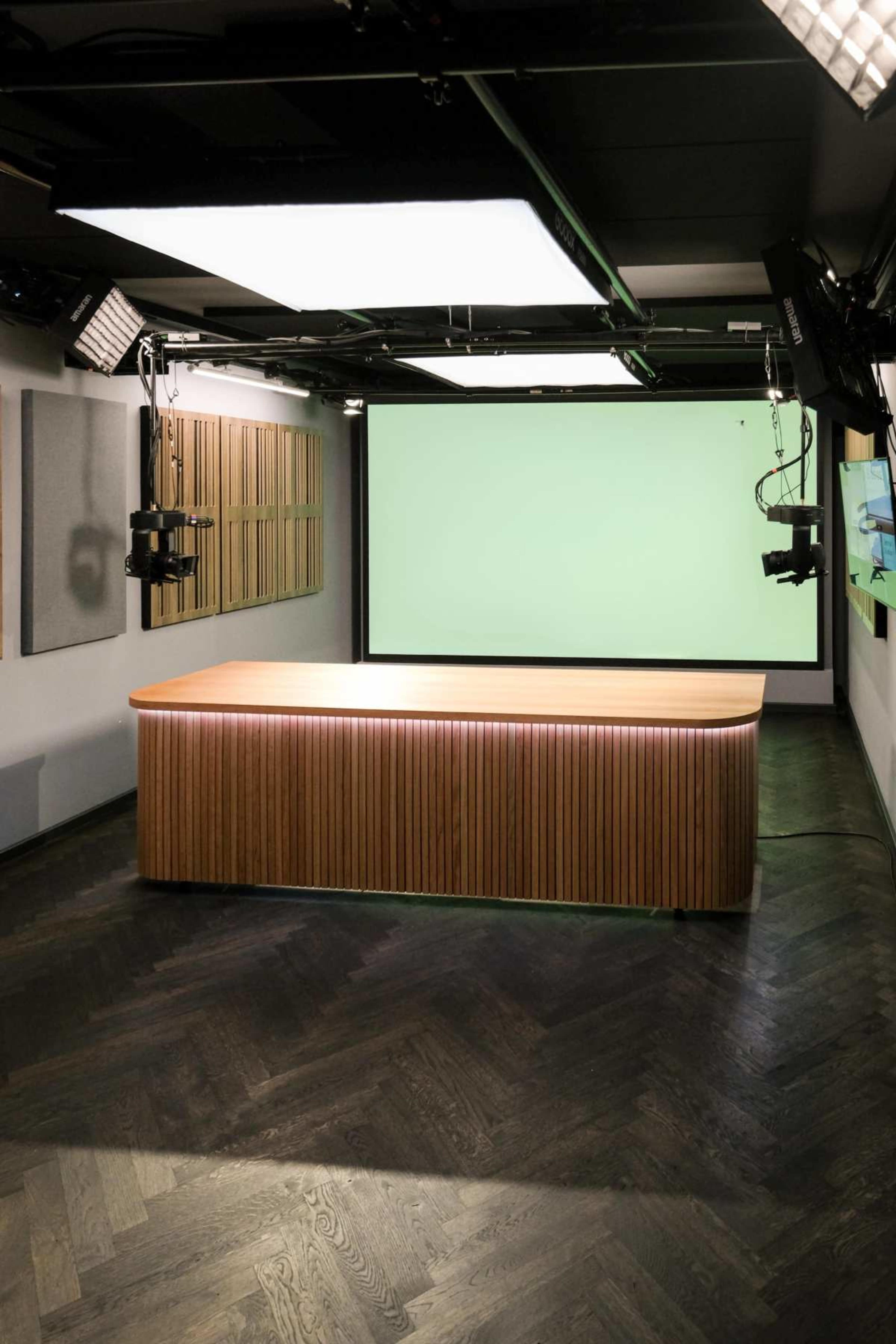 The image shows a modern studio with a green screen backdrop, wooden table at the center, and numerous studio lights mounted on the ceiling.