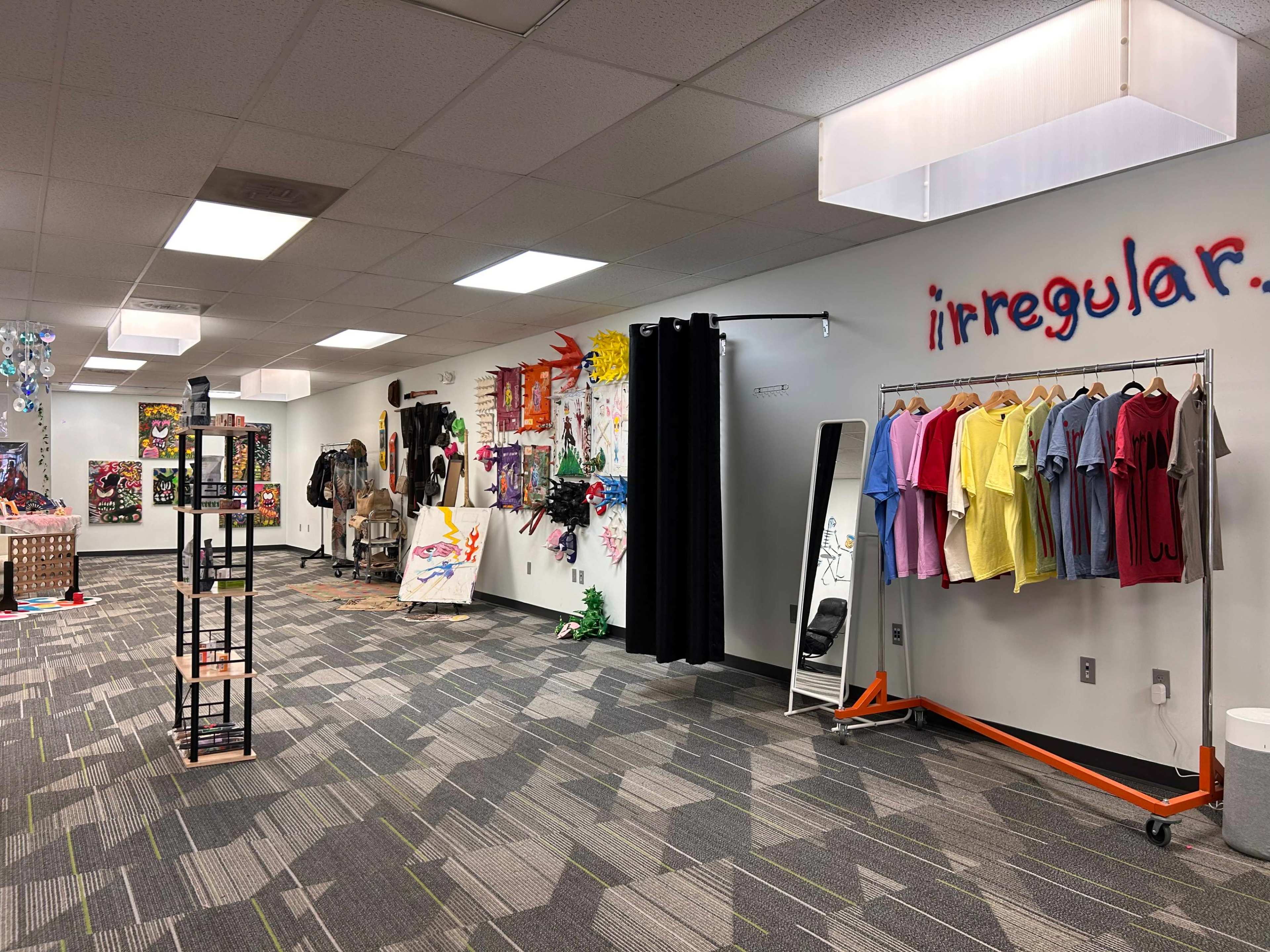The image shows a spacious room filled with colorful artworks on the walls, racks of clothing, and different display items, suggesting a creative workspace or art studio.