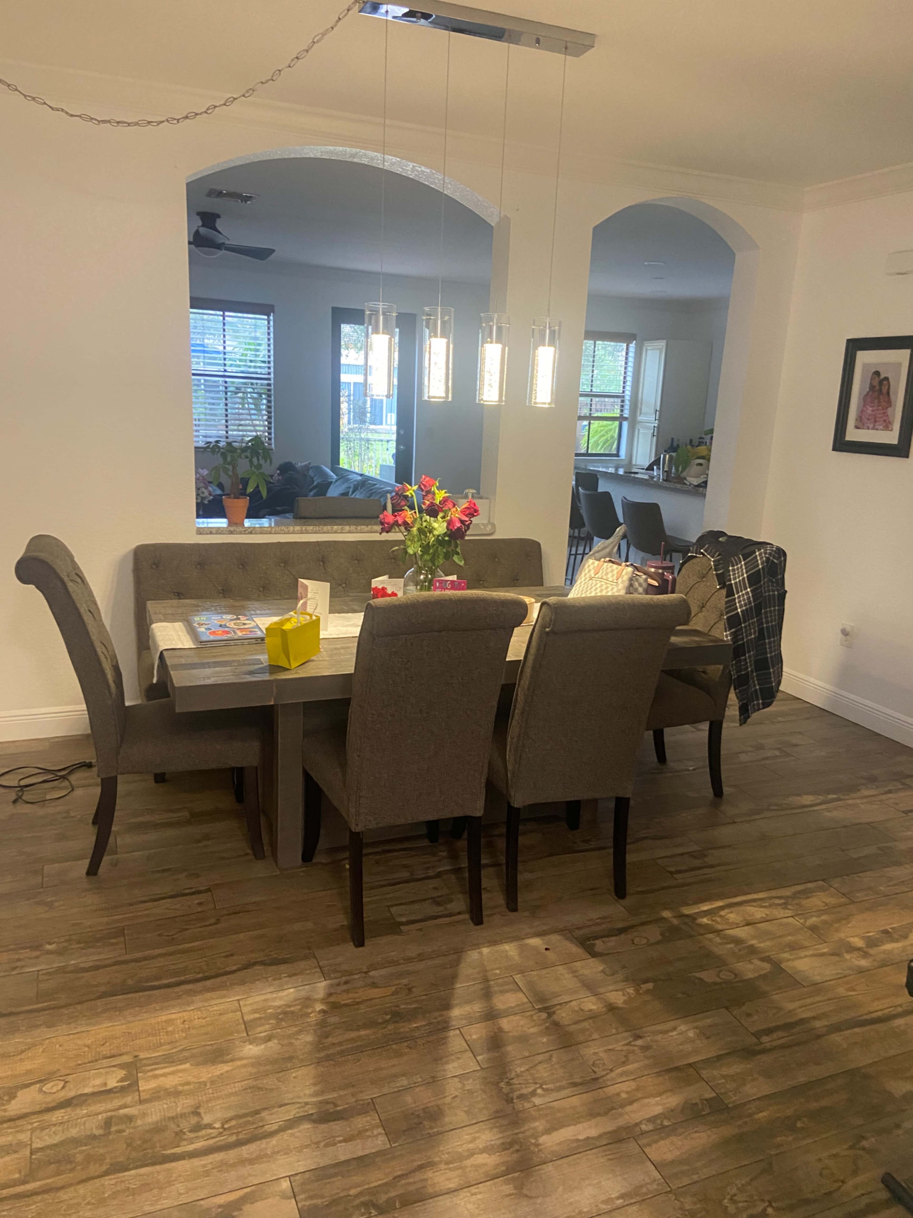 A dining area features a rectangular table with gray chairs, a vase of flowers, and modern pendant lights hanging above it, adjacent to a spacious living area with a sofa and large windows.