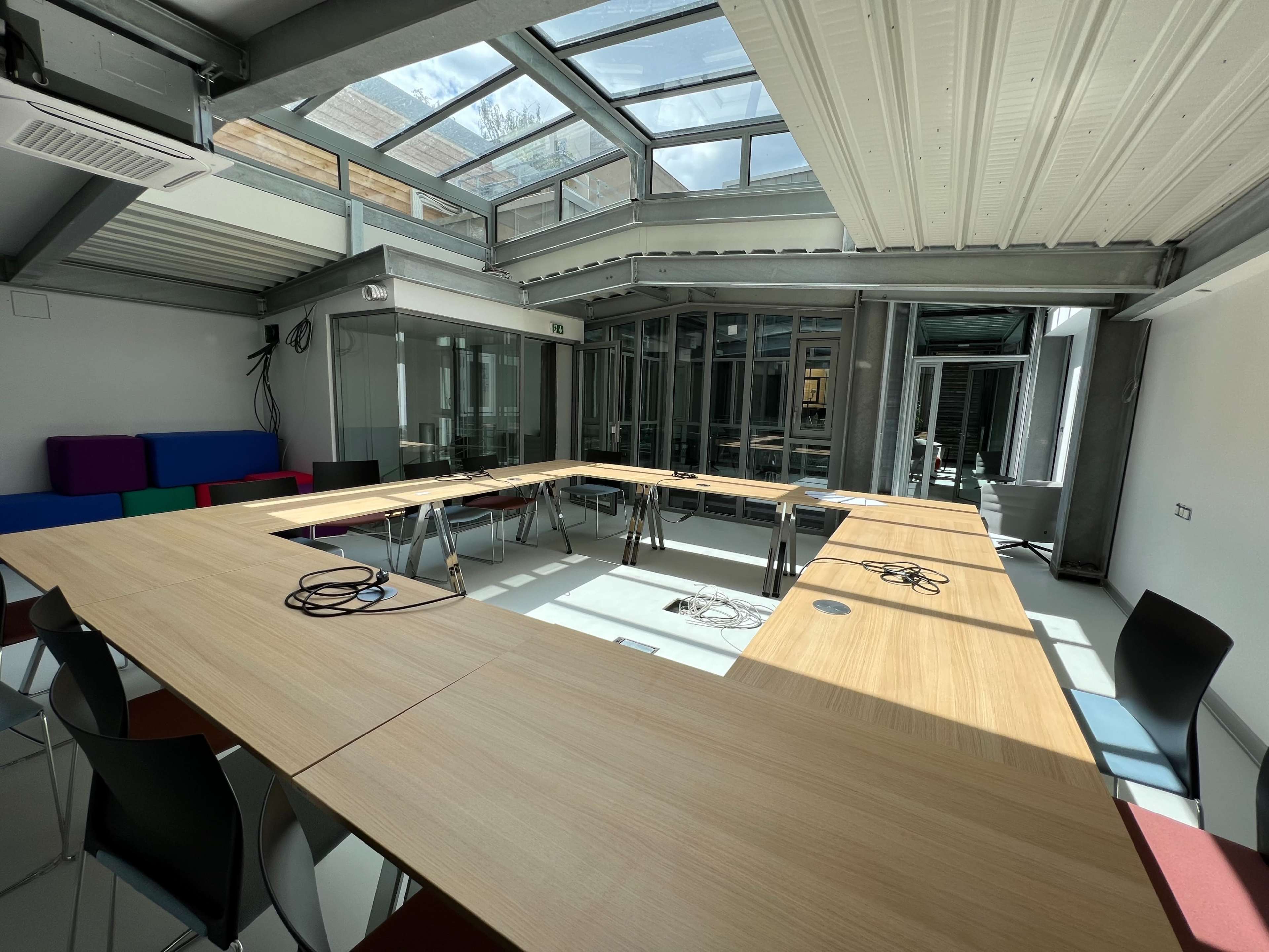A large, modern conference room features a rectangular arrangement of tables, surrounded by chairs, with a glass roof allowing natural light to fill the space.