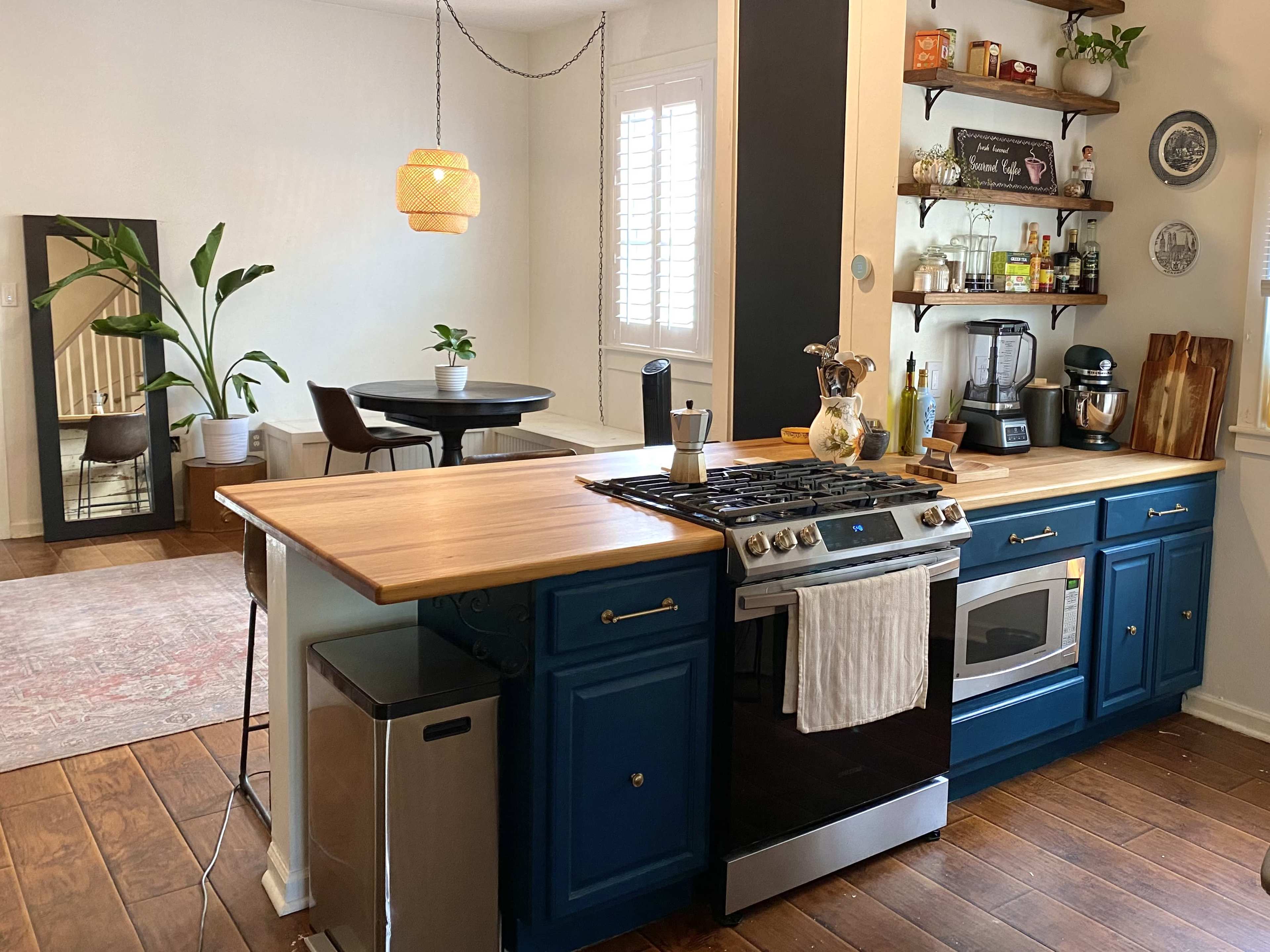 The image depicts a modern kitchen featuring blue cabinets, a gas stove, and a wooden countertop, with an adjoining dining area visible in the background.