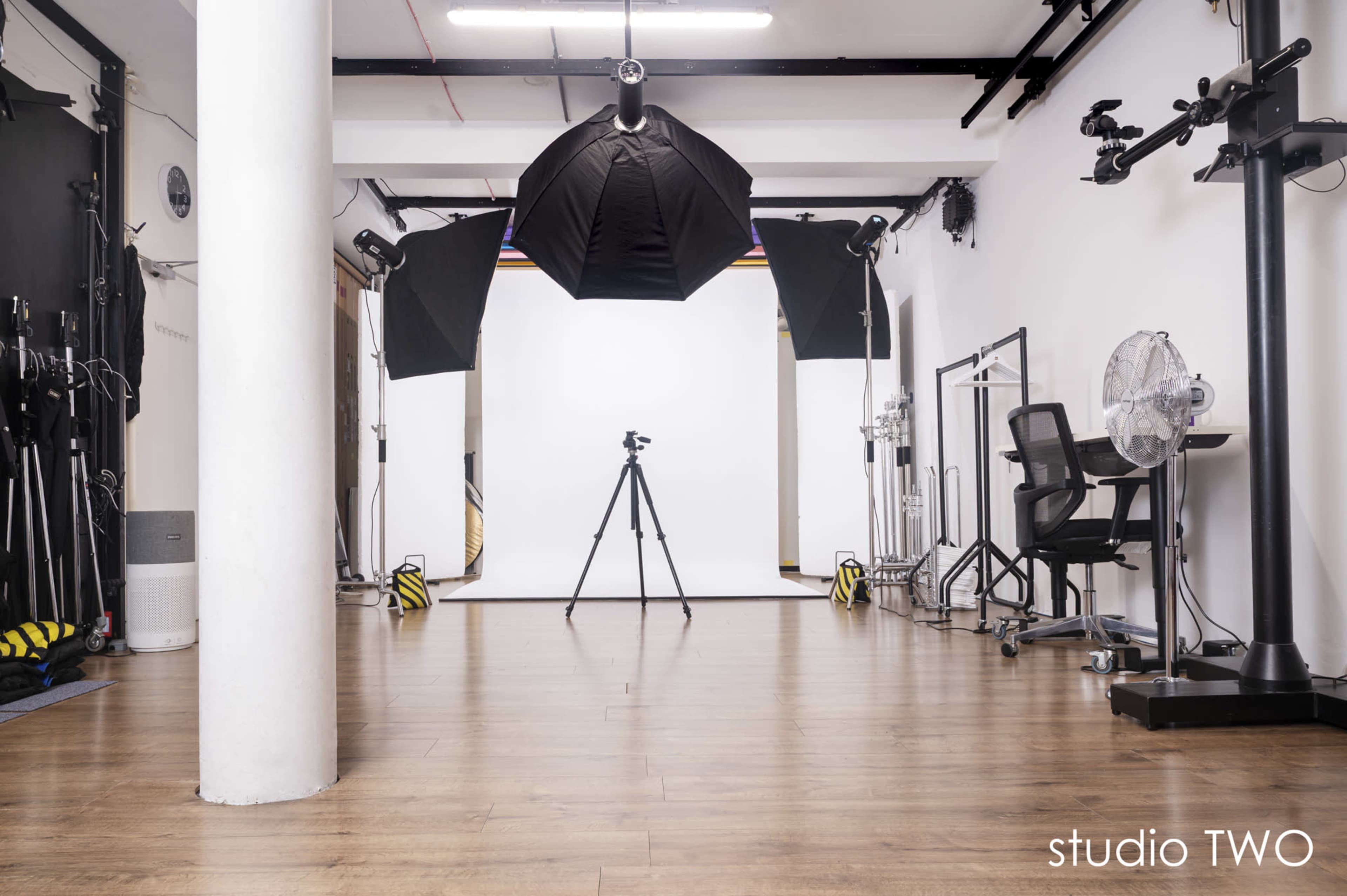 The image shows a photography studio with a white backdrop, lighting equipment on either side, a tripod positioned in the center, and a chair in the corner.