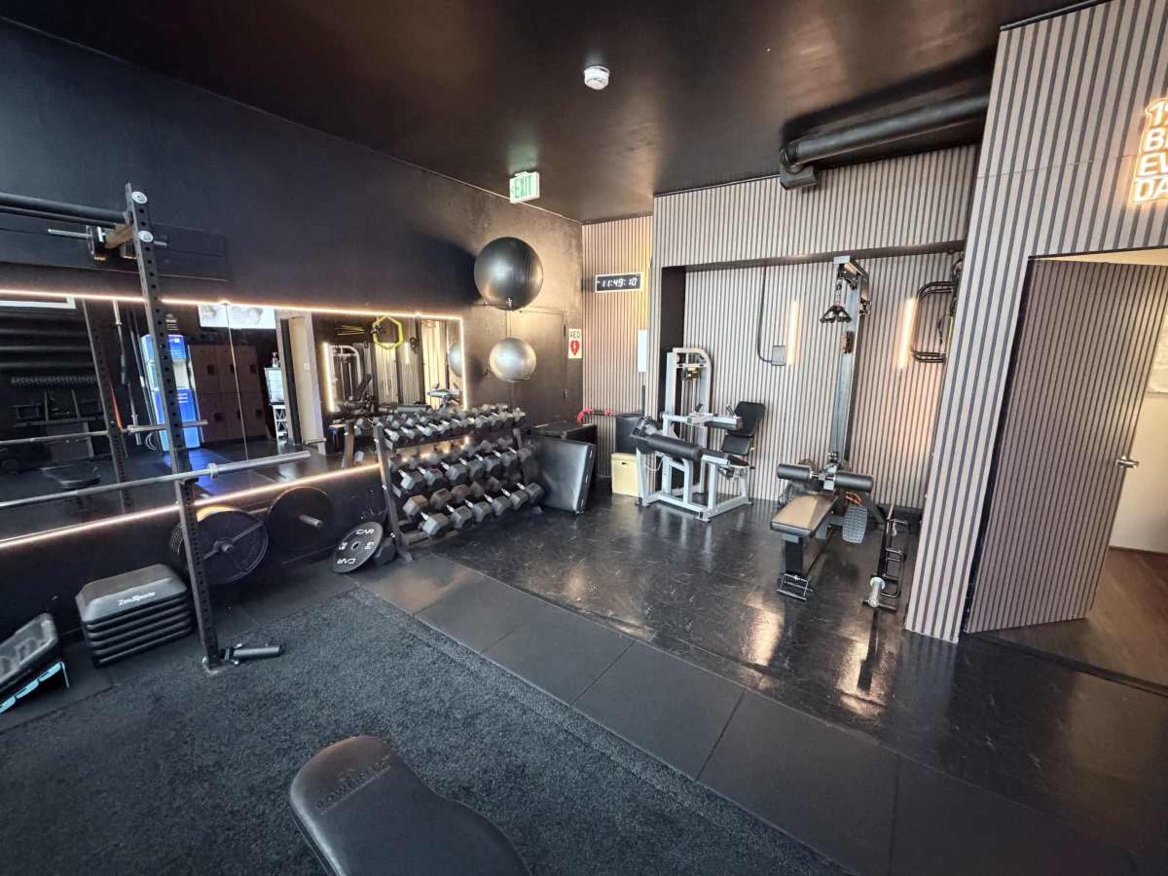 The image shows a modern gym interior featuring various workout equipment, weights, and exercise balls arranged neatly in a dimly lit space with black walls.