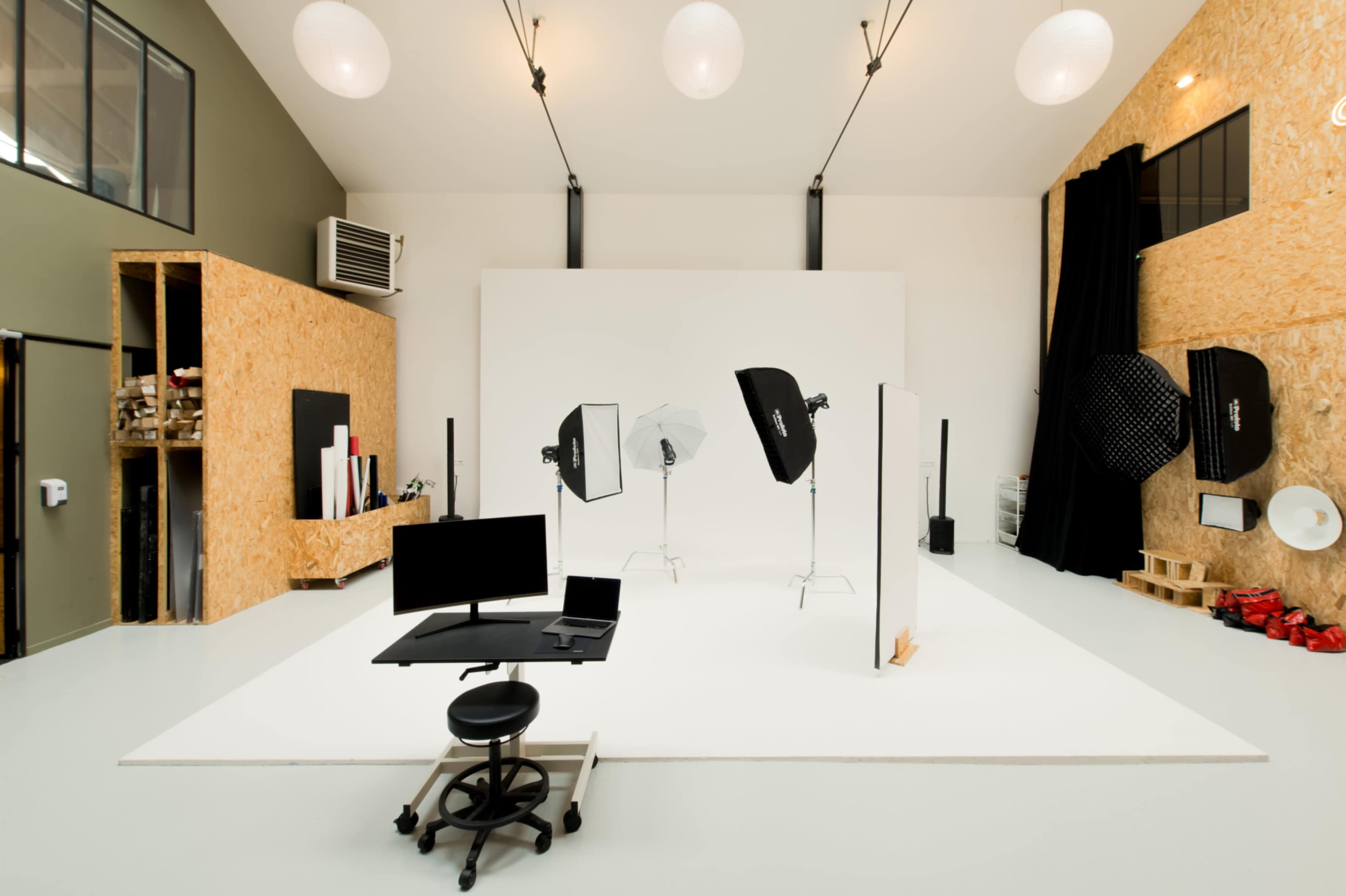 A photography studio with a white backdrop, lighting equipment, a workstation, and various props organized along the walls.