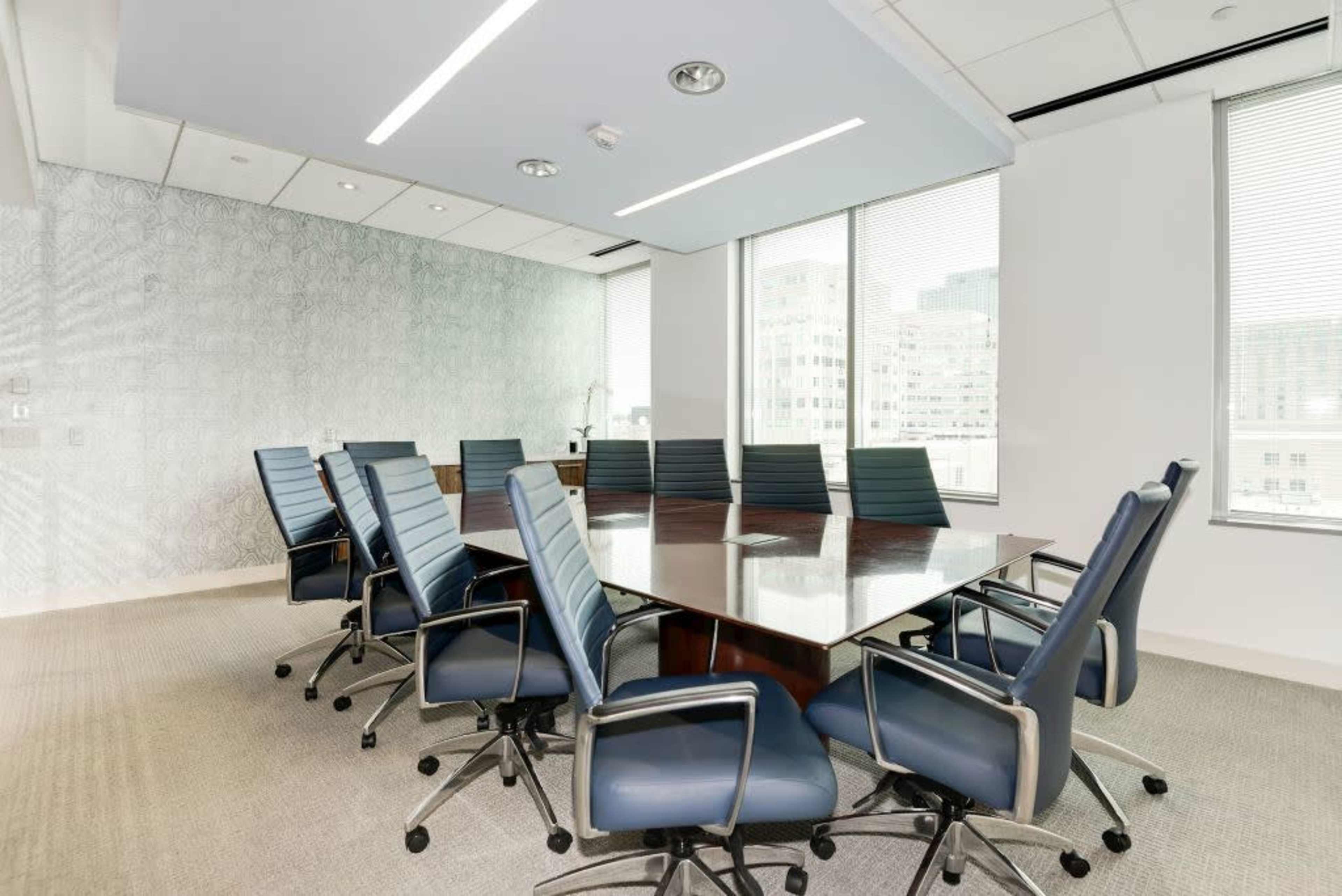 The image shows a modern conference room with a large rectangular table surrounded by blue office chairs, and windows providing a view of a cityscape.