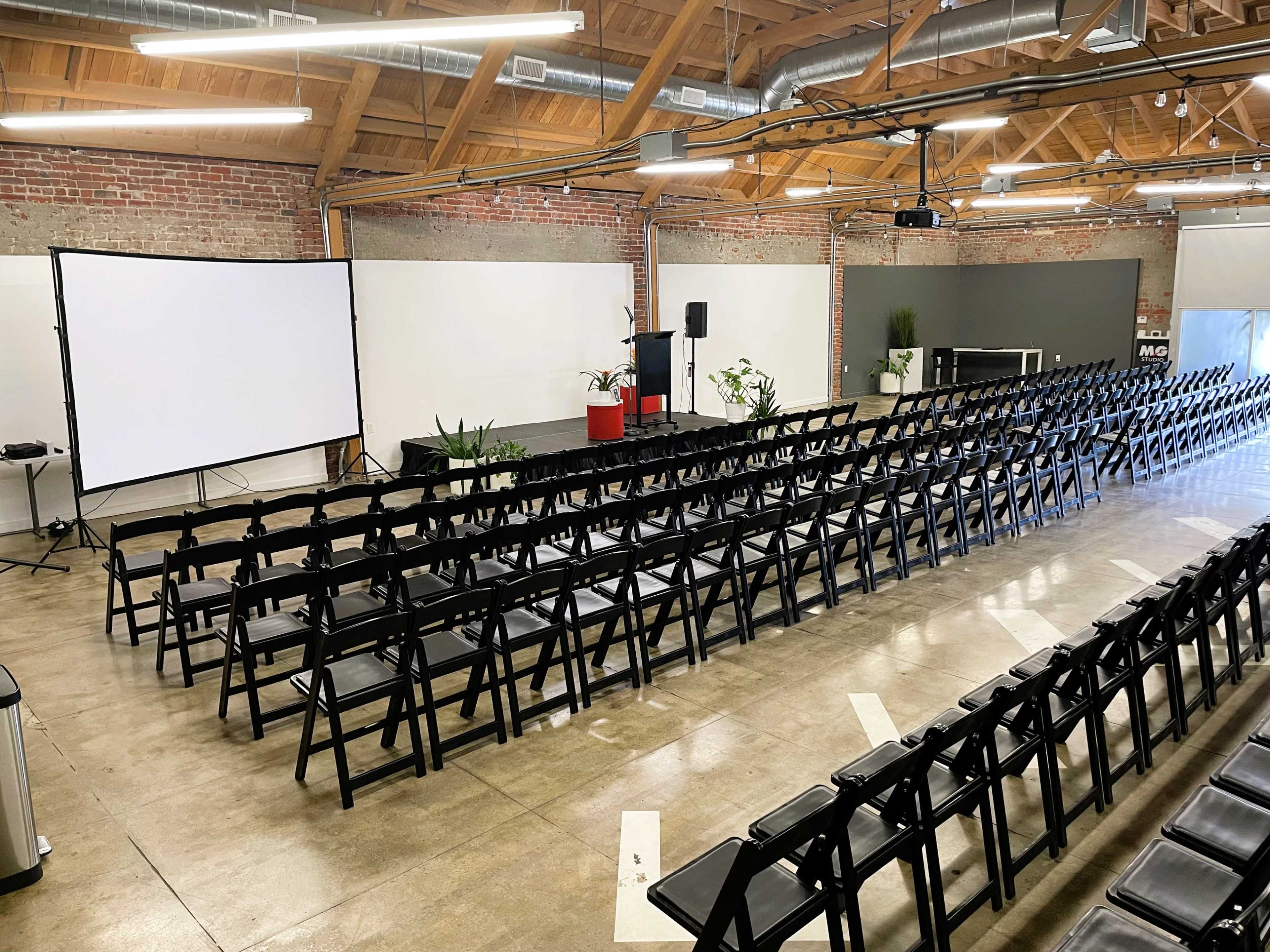 The image shows a spacious room arranged with rows of black folding chairs facing a large projection screen and a podium, with exposed brick walls and a wooden ceiling.
