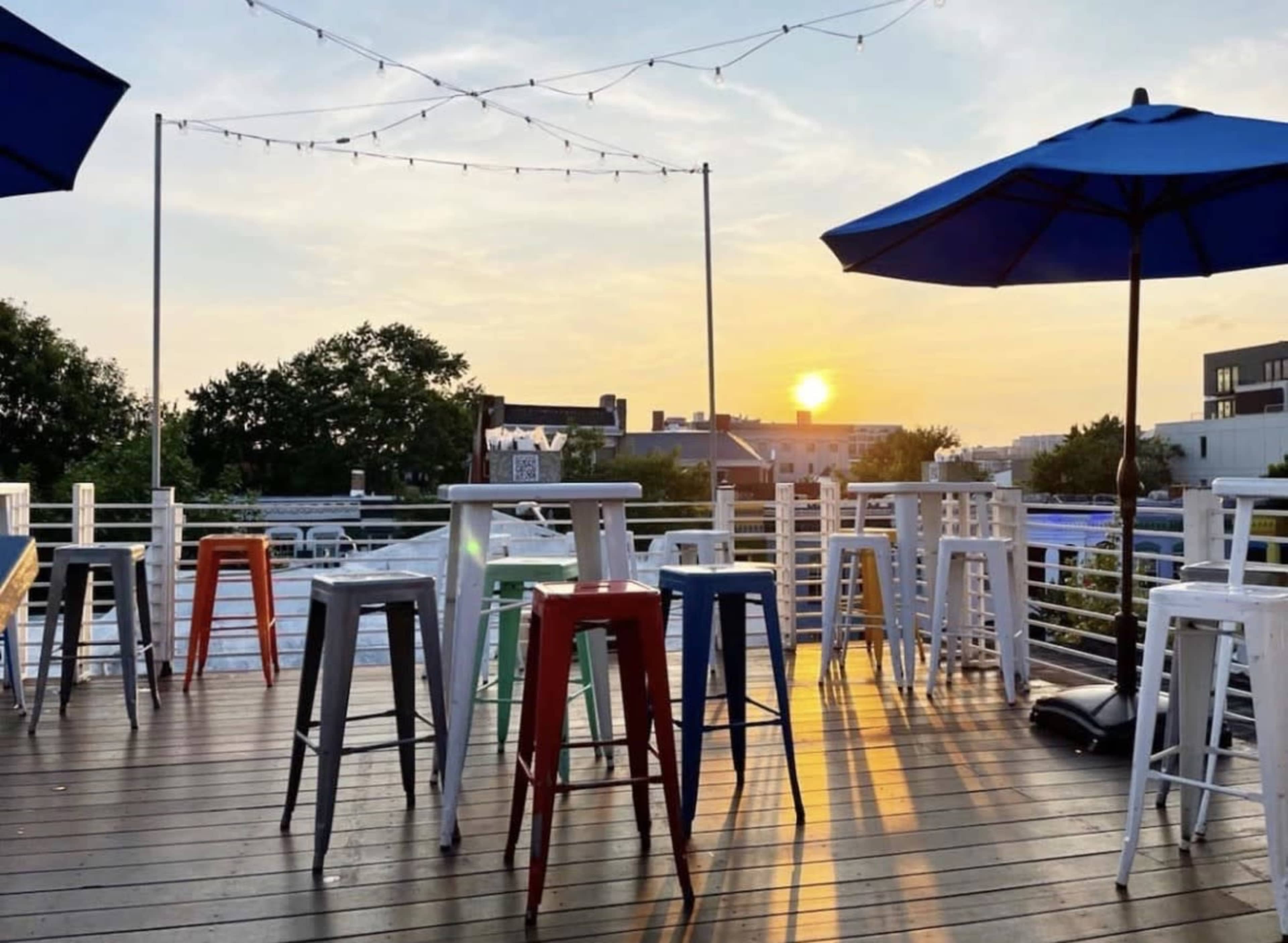 A rooftop terrace features several tall stools and tables under blue umbrellas with a sunset in the background.