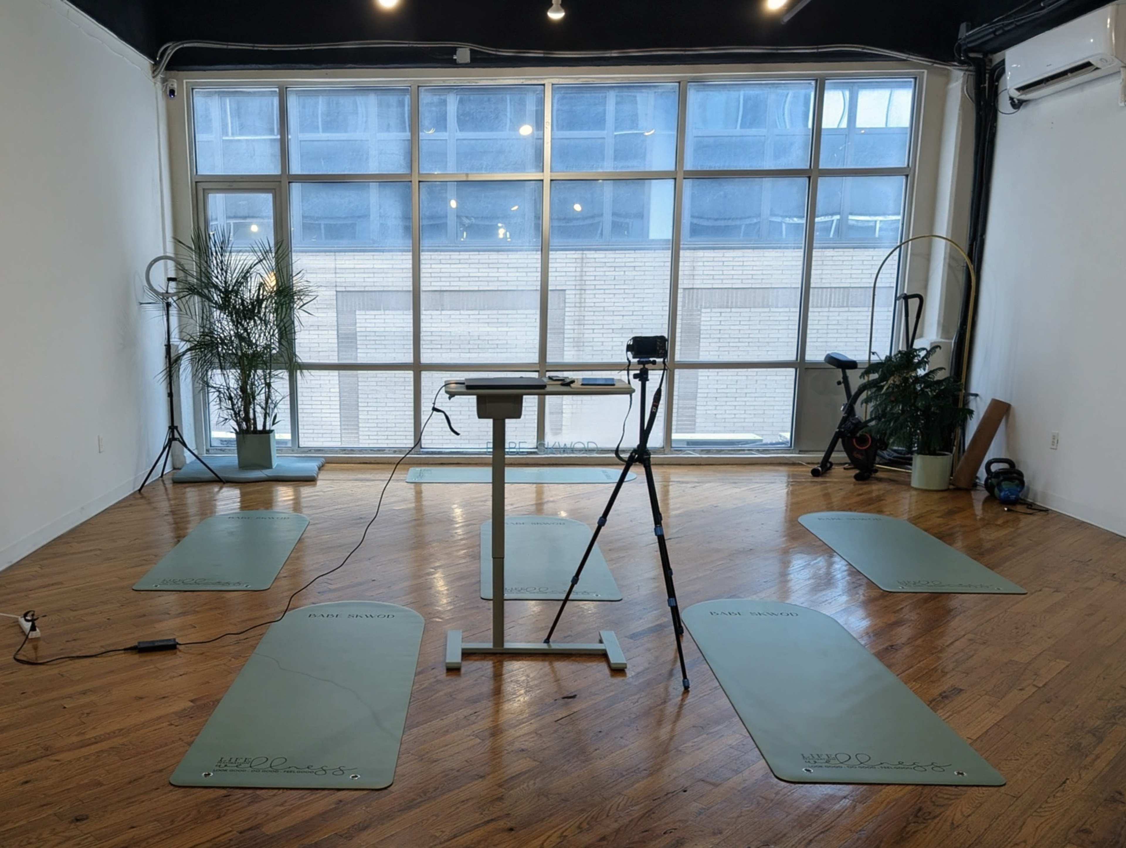The image shows a spacious studio with wooden flooring, six yoga mats arranged on the ground, and a camera setup facing the mats.