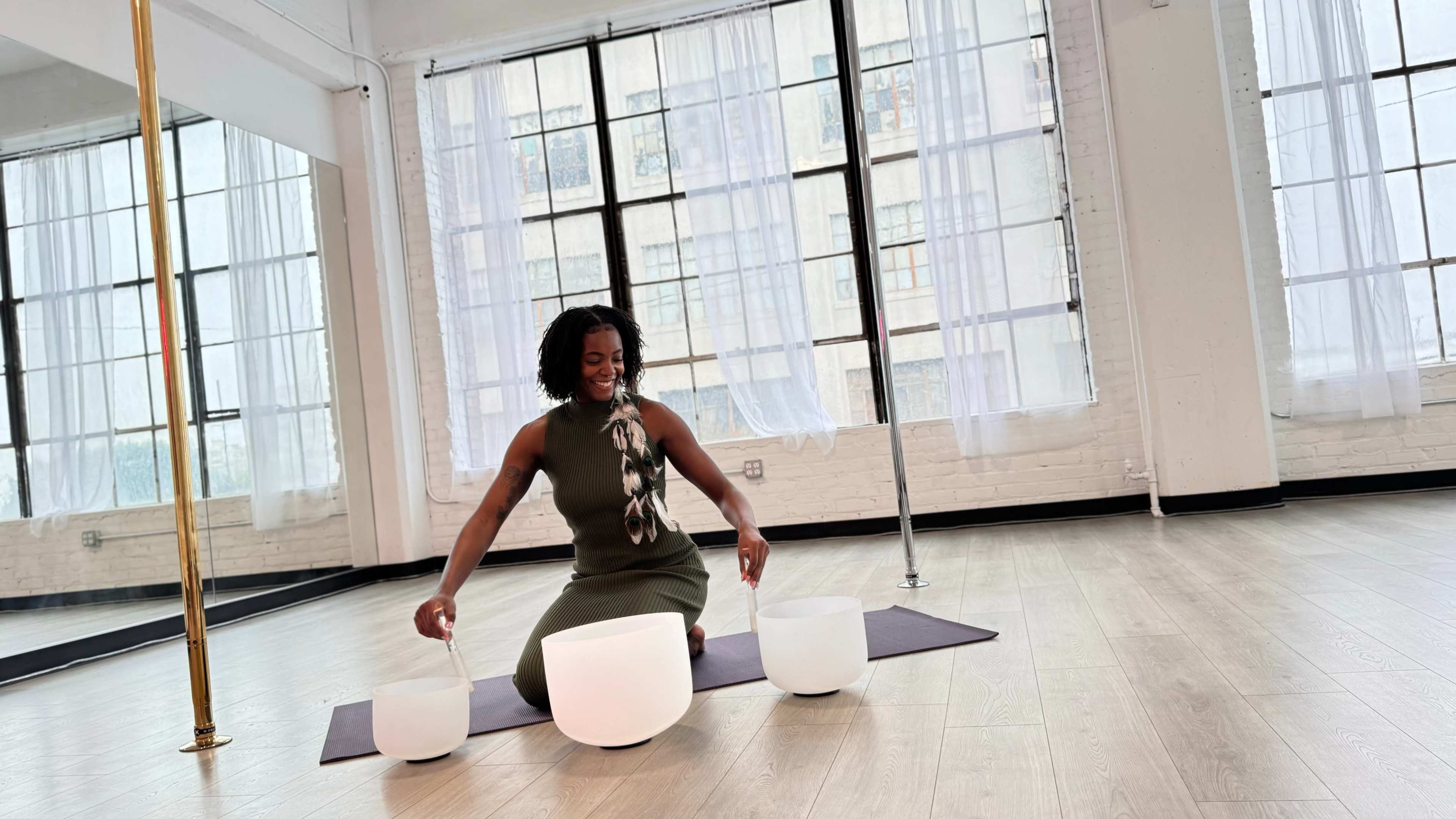A woman sits on a yoga mat in a room with large windows, positioned between two white bowls.