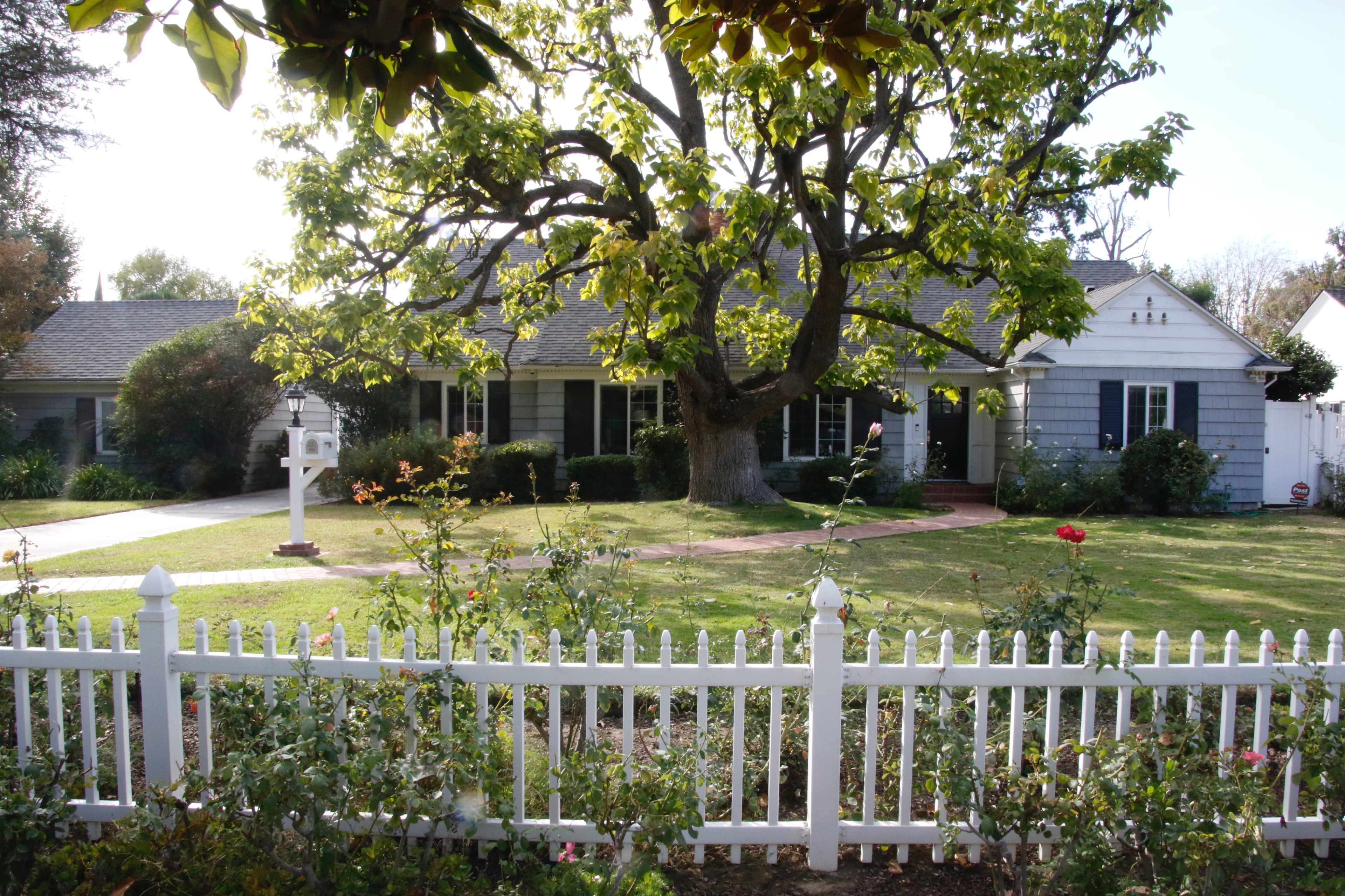 A single-story house with a gray exterior and a large tree in the front yard is surrounded by a white picket fence and well-maintained landscaping.
