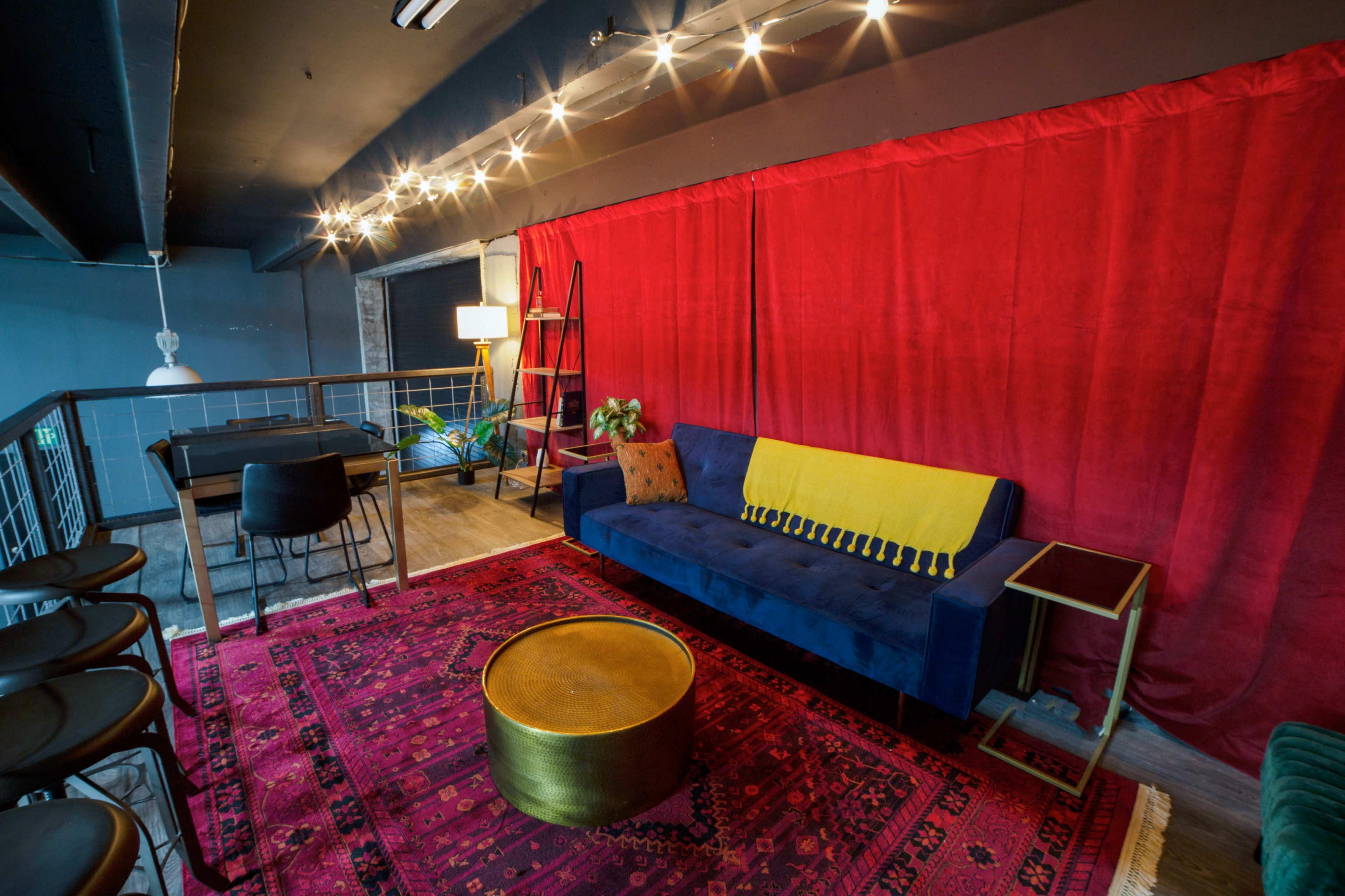 The image shows a modern lounge area featuring a blue velvet sofa with a yellow throw, a round gold coffee table, a red curtain backdrop, and various plants and furnishings.