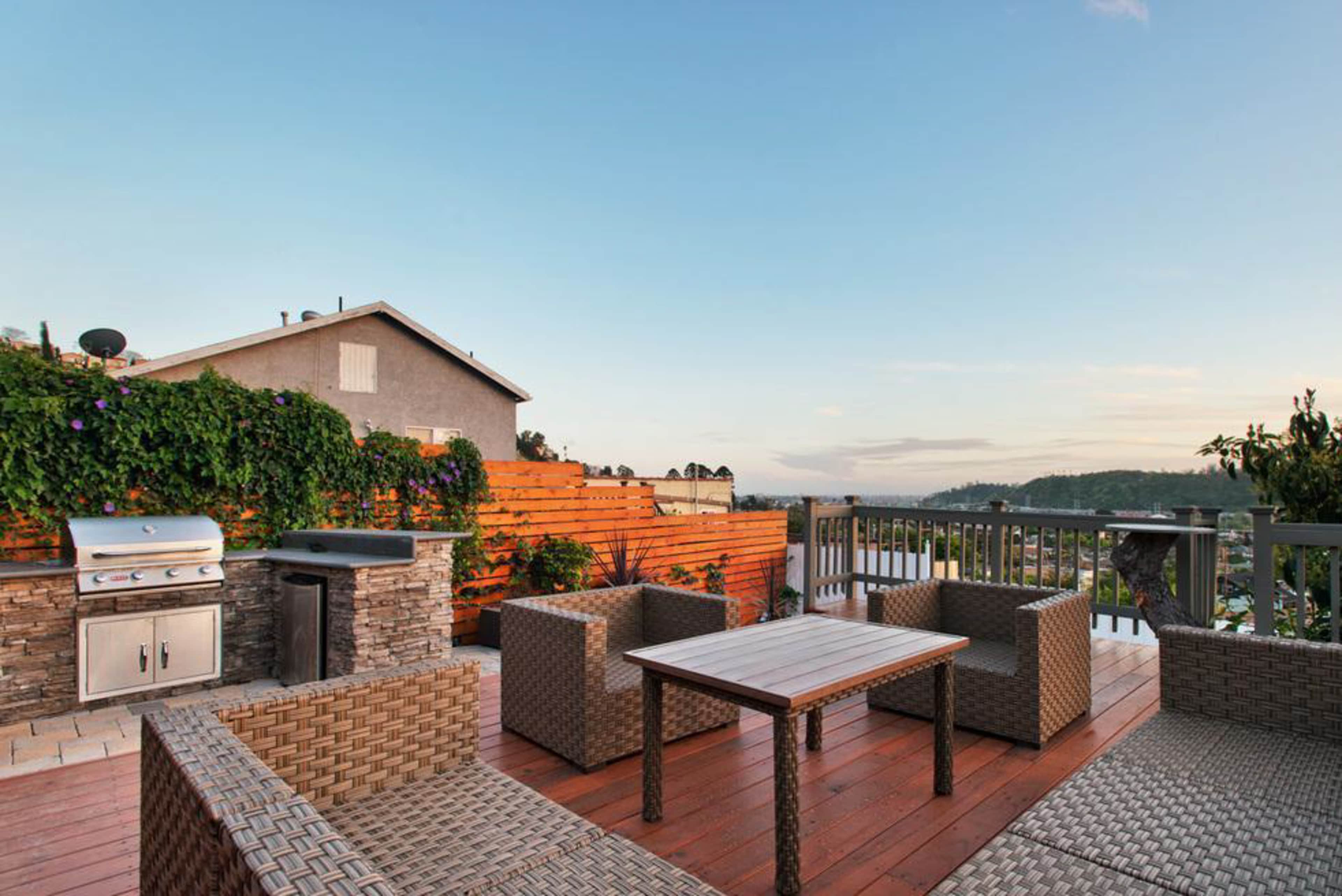A wooden deck with a table and chairs, a grill, and a hillside view in the background.