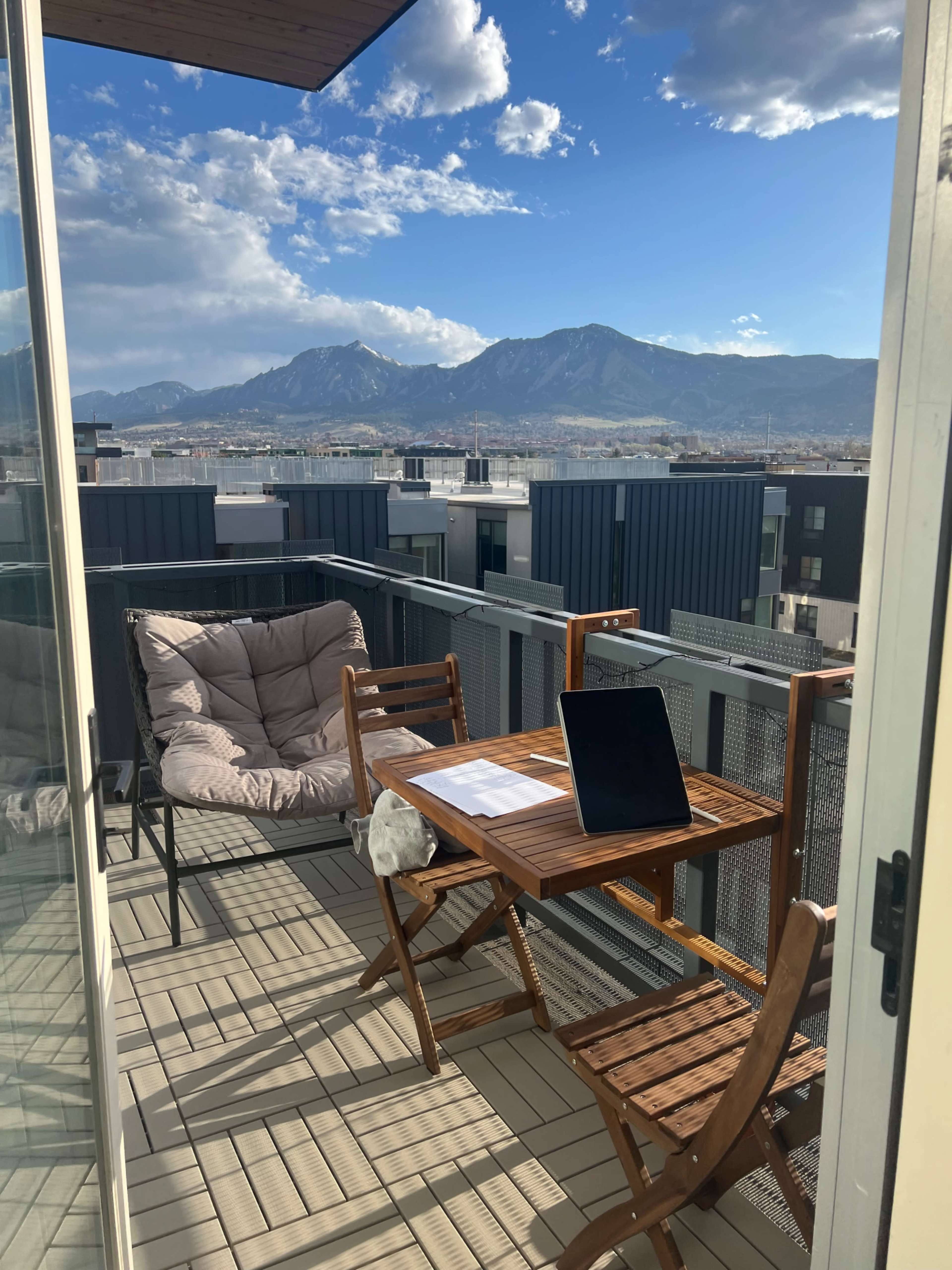 A balcony features a wooden table with a laptop and papers, surrounded by two chairs, overlooking a mountainous landscape under a partly cloudy sky.