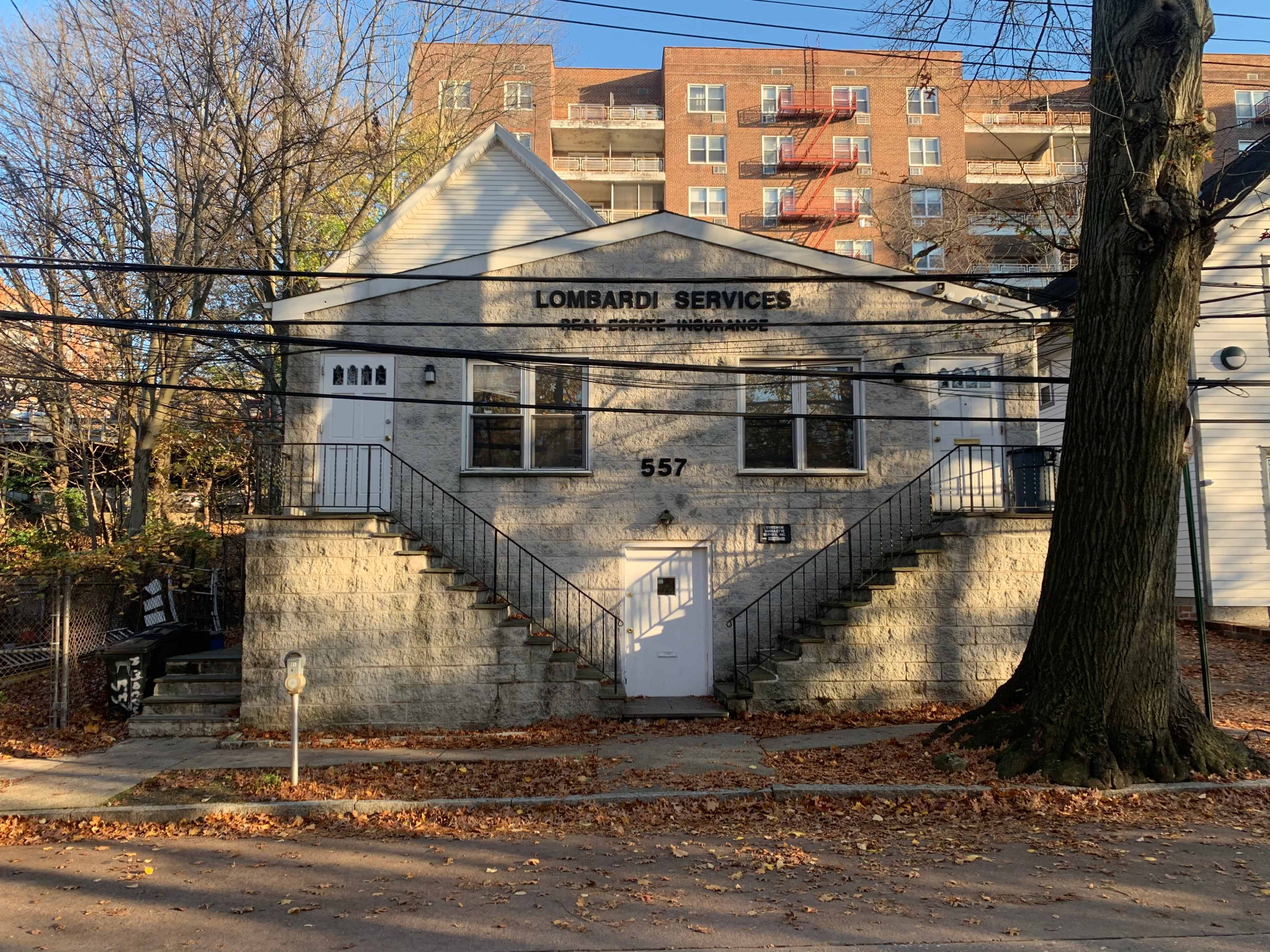 A two-story stone building with the name "Lombardi Services" displayed above its entrance, featuring a staircase on each side and surrounded by autumn foliage and power lines.