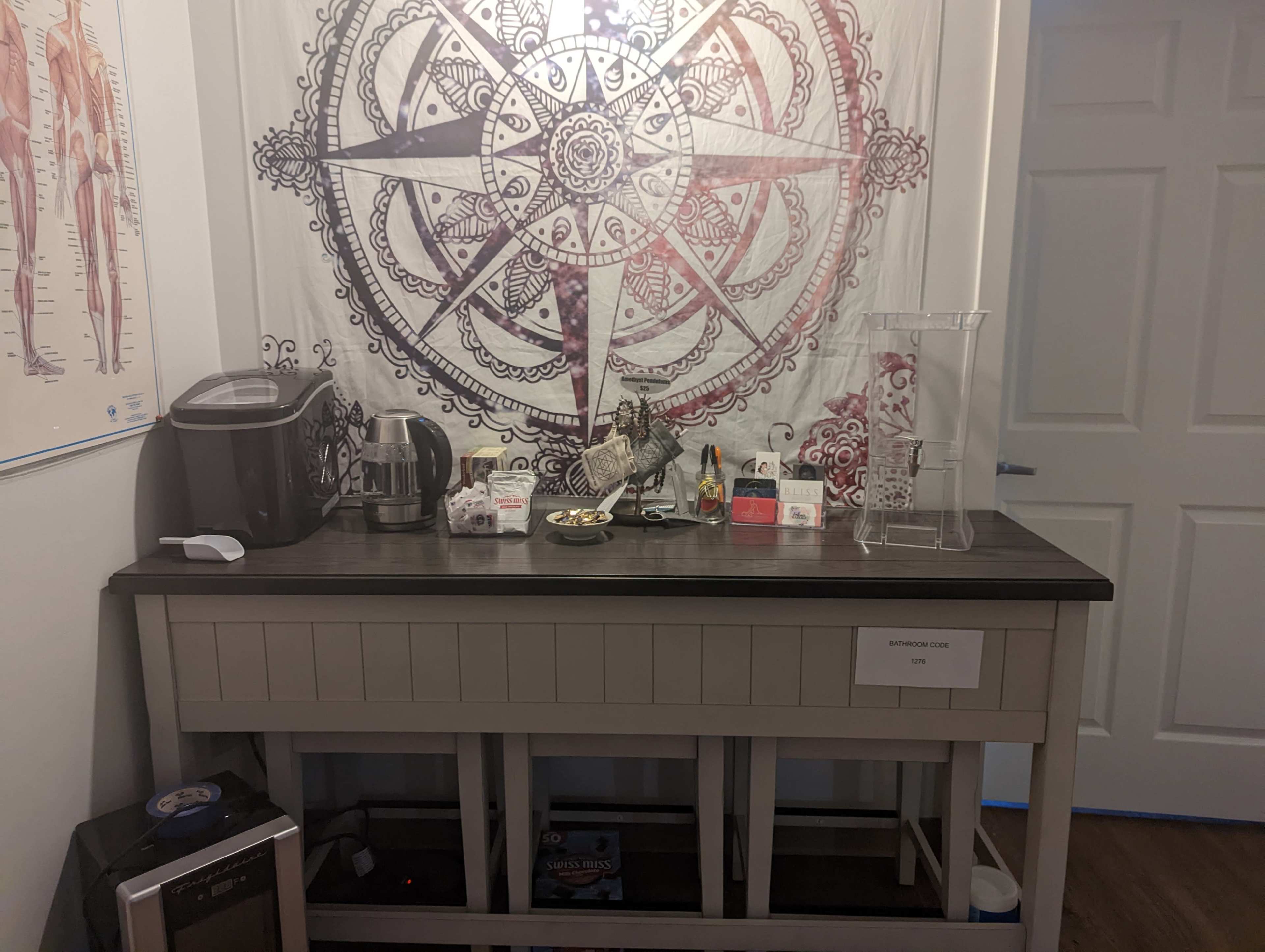 A room corner with a table displaying a coffee machine, toaster, snacks, and decorative items against a wall featuring a large mandala tapestry.
