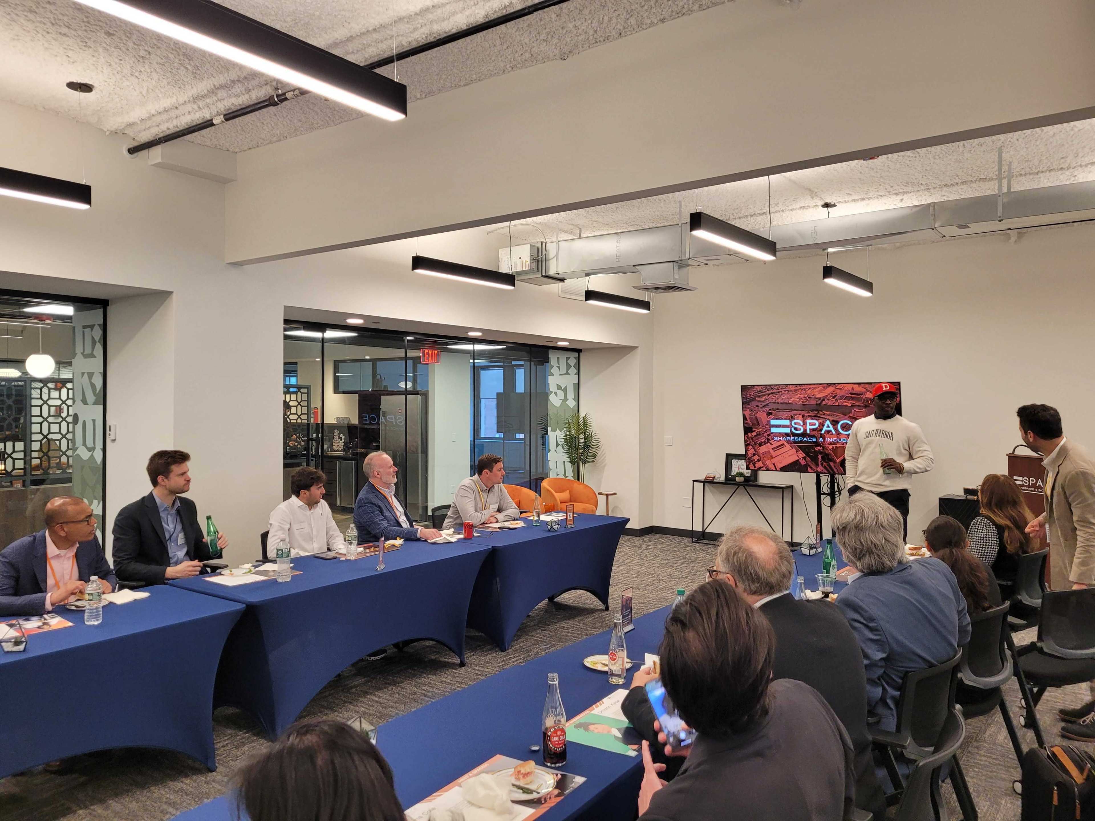 A group of professionals is gathered around a large table in a modern conference room, watching a presentation displayed on a screen at the front.