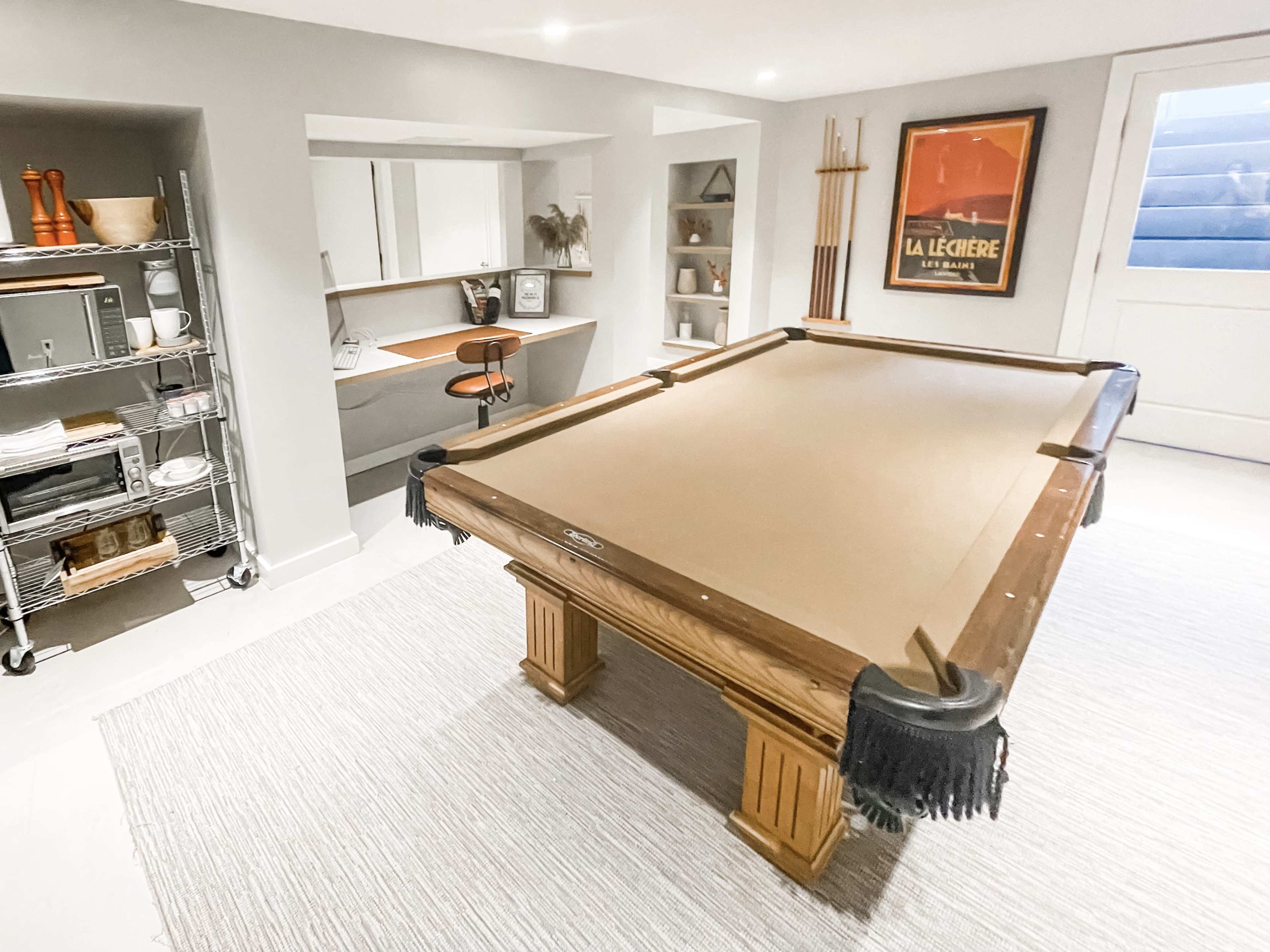 The image shows a basement game room featuring a large pool table, a sleek bar area, and organized shelving with various items.