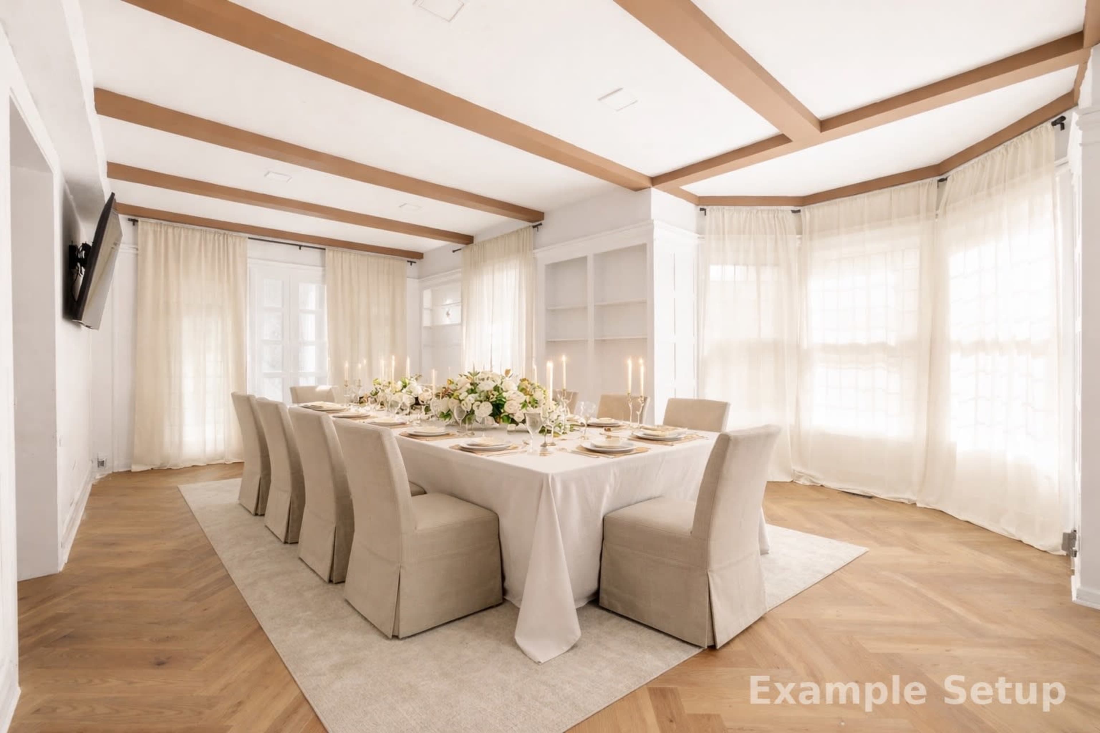 A long dining table is set with elegant tableware and floral centerpieces in a bright room with large windows and light-colored curtains.