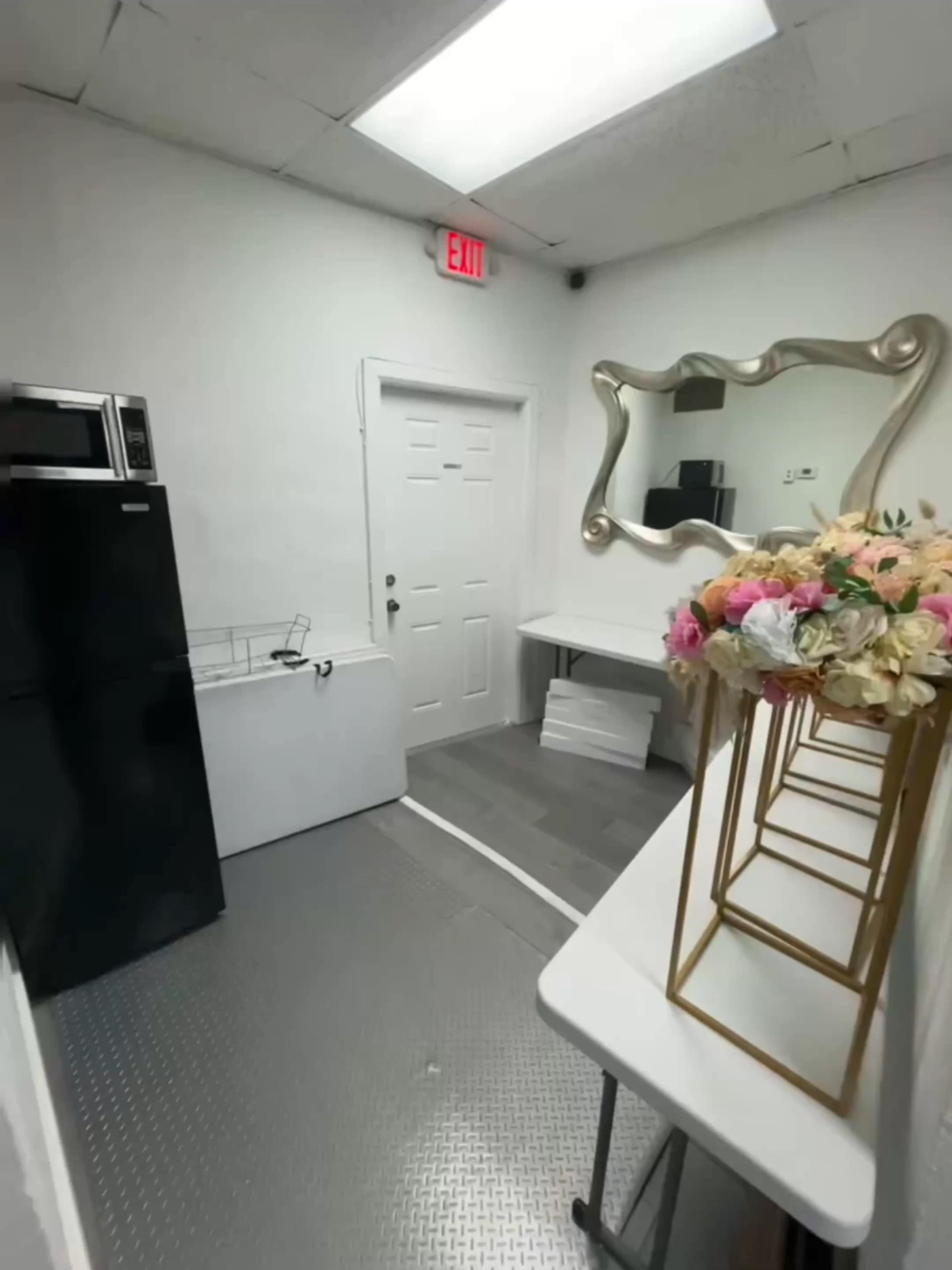 The image shows a small room with a black refrigerator, a white door, a large mirror, and a table displaying a floral arrangement.