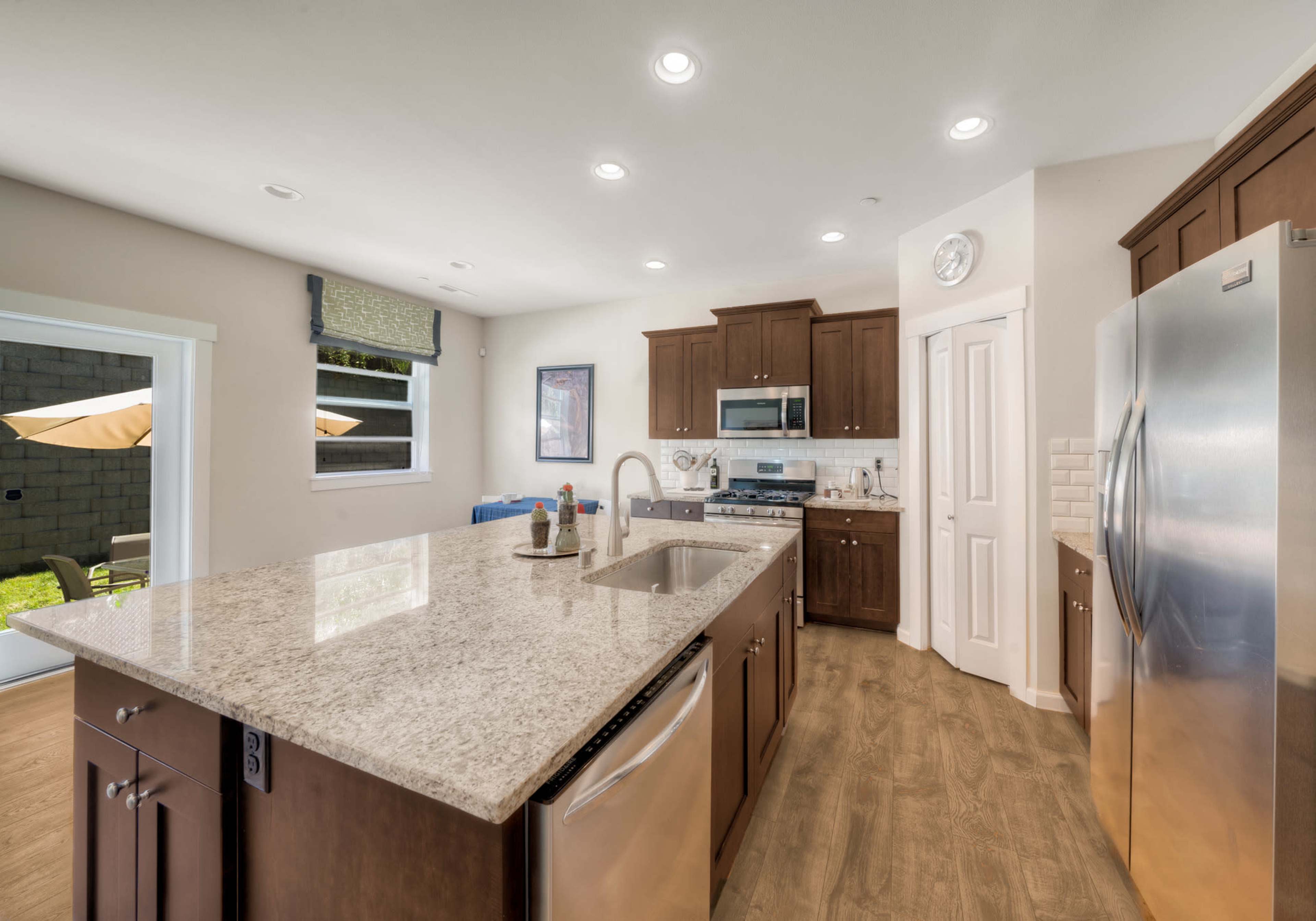 A modern kitchen features wooden cabinets, a large granite countertop island, stainless steel appliances, and a window overlooking an outdoor space.
