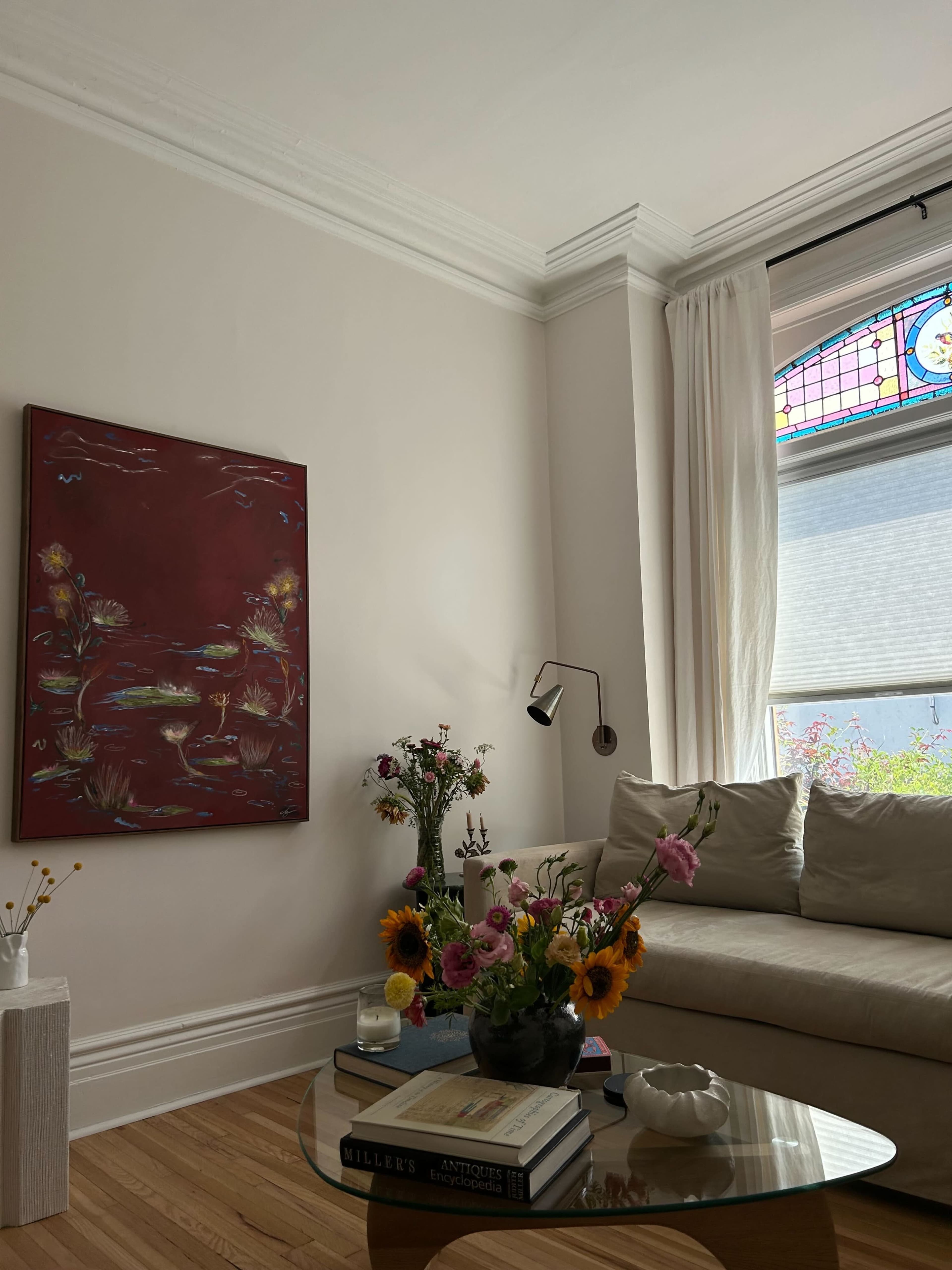 A cozy living room features a colorful painting on the wall, a beige couch, a round glass table adorned with a floral arrangement, and a window with a stained glass design.