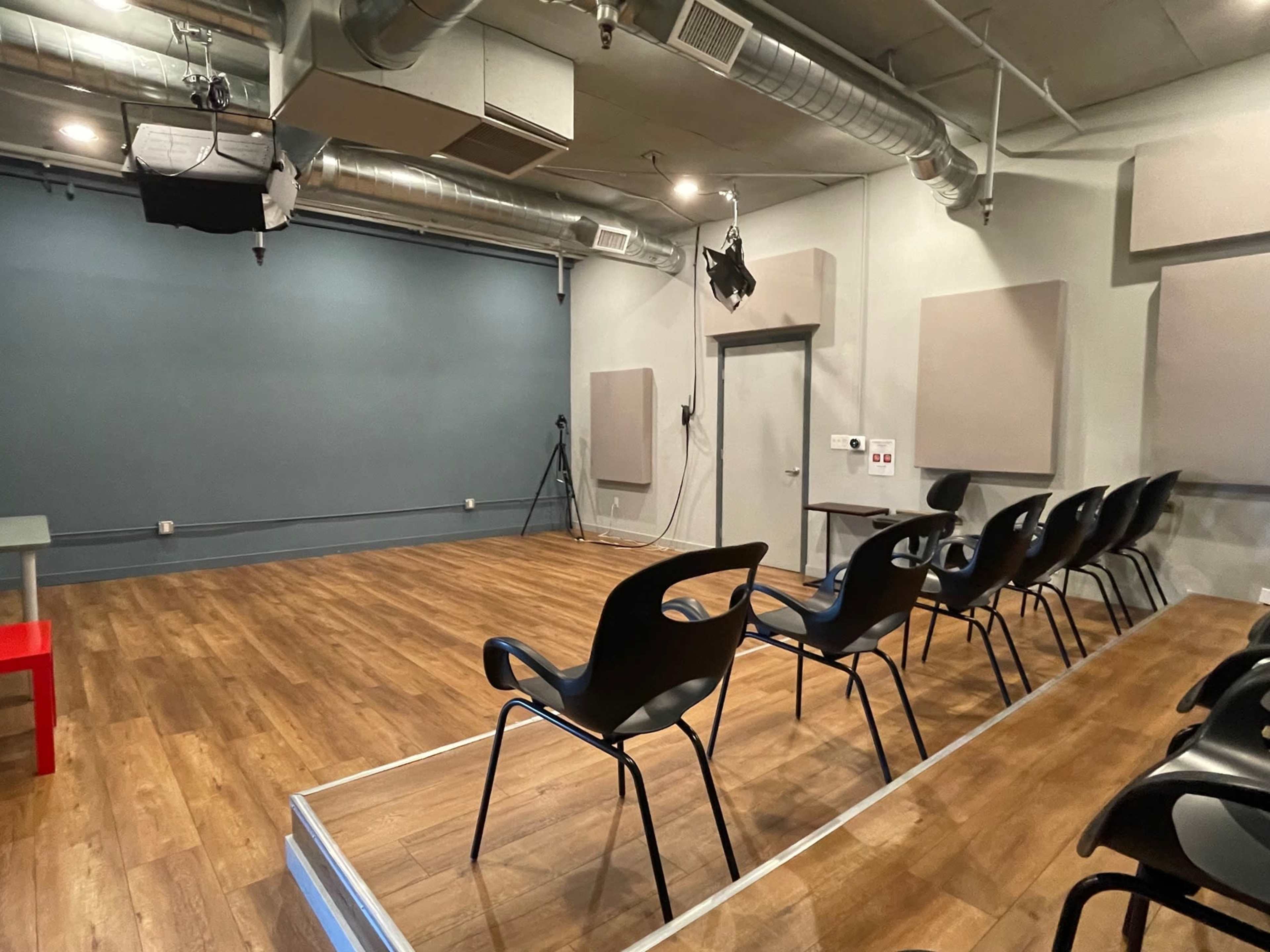 A small performance space with wooden flooring, featuring a row of black chairs facing a gray wall, and acoustic panels on either side.