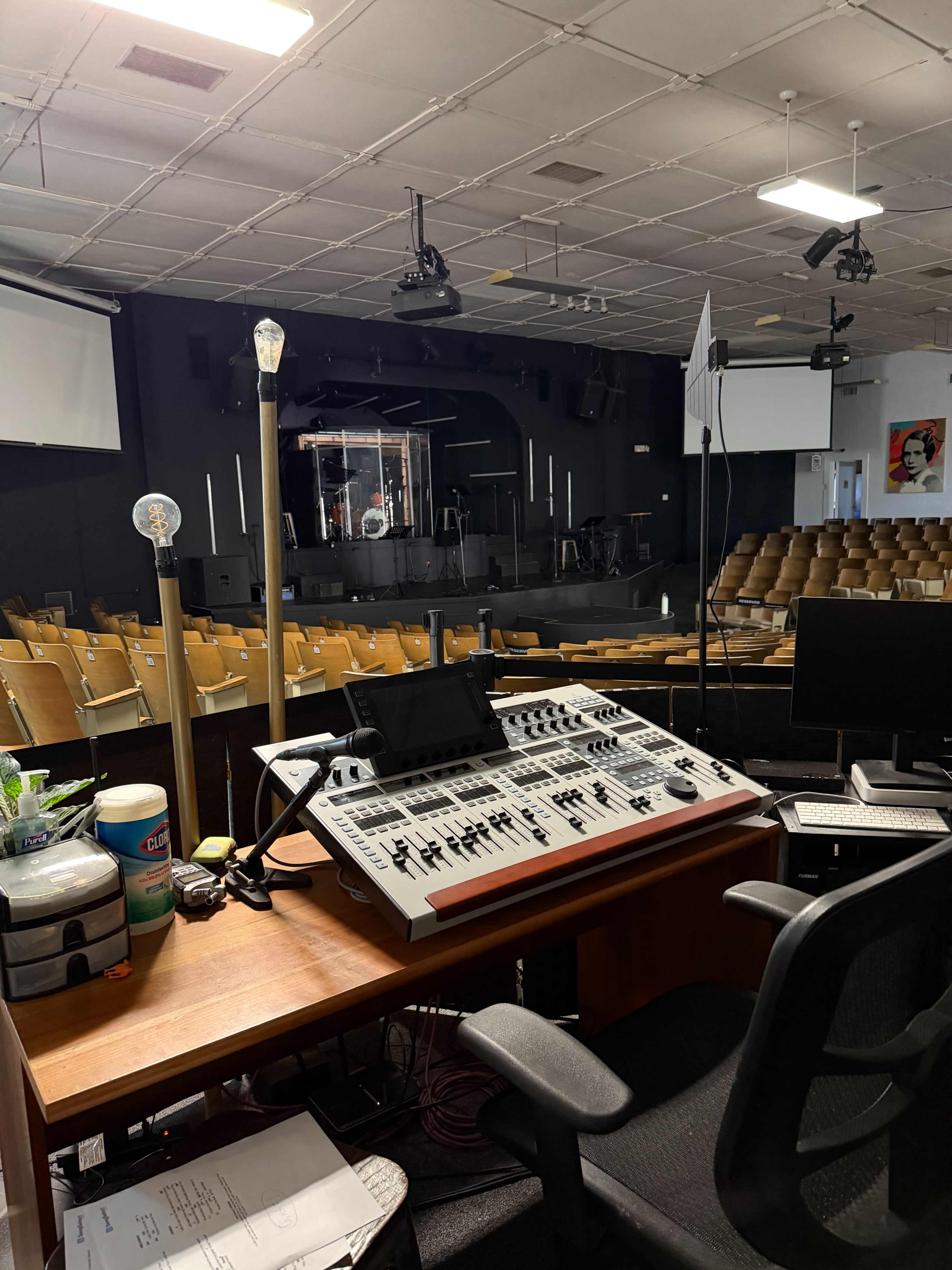 The image shows a sound control station in a concert hall with empty seating and a stage set for a performance.