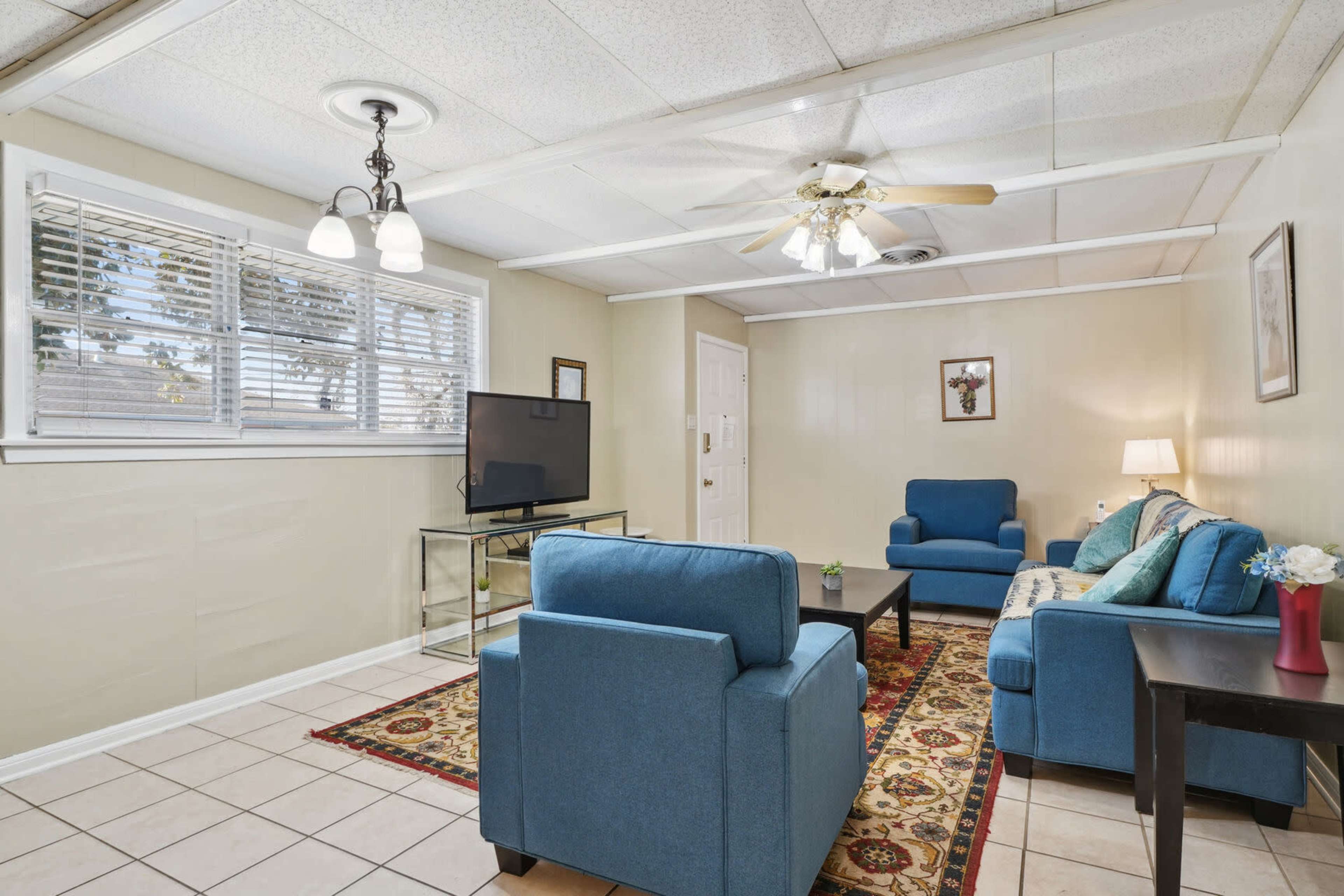 A living room features two blue chairs, a sofa with decorative cushions, a glass coffee table, and a television on a stand, with large windows allowing natural light.